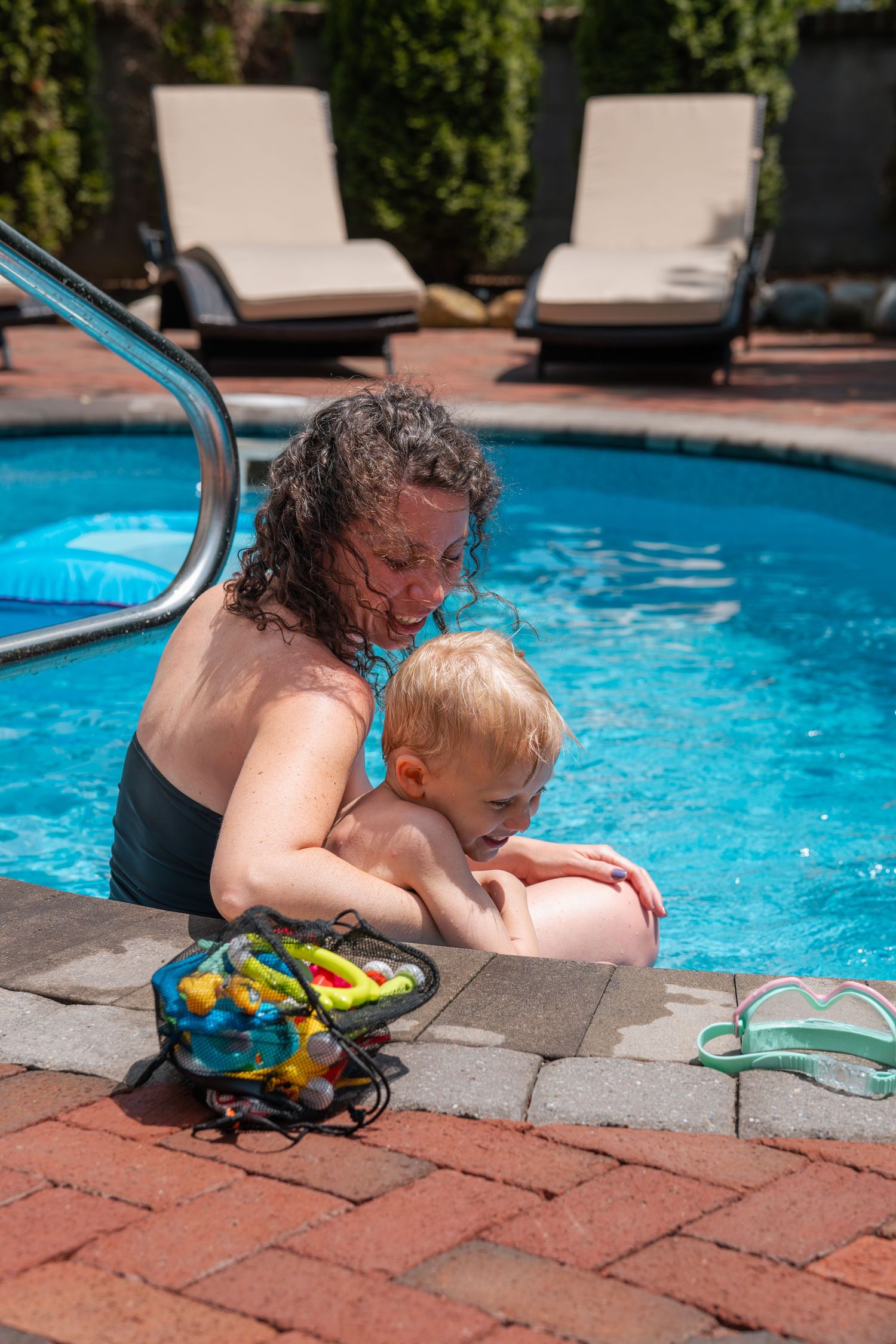 Mother holding toddler at pool edge in backyard swimming pool with lounge chairs in background on sunny day.
