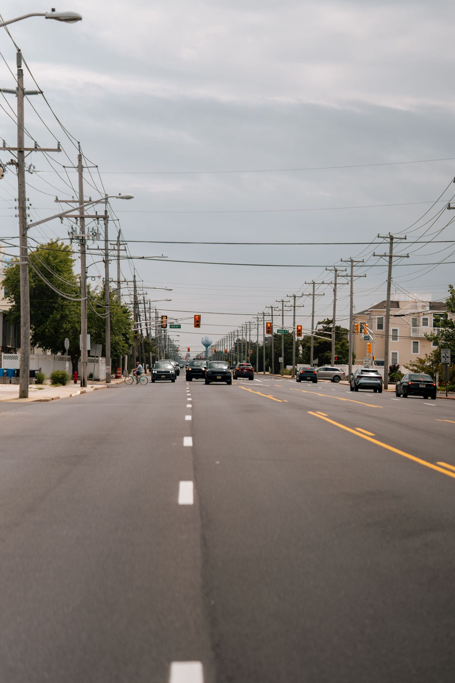 A wide empty road with utility poles, traffic lights, and distant vehicles under a cloudy sky in a suburban area.