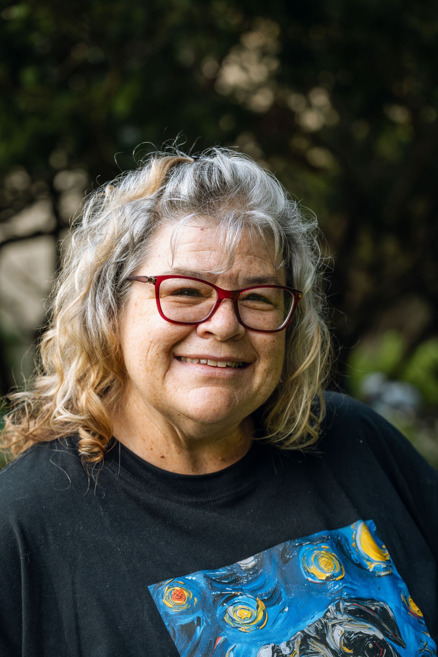 Smiling older woman with curly gray hair and red glasses wearing a black shirt with colorful art print outdoors.