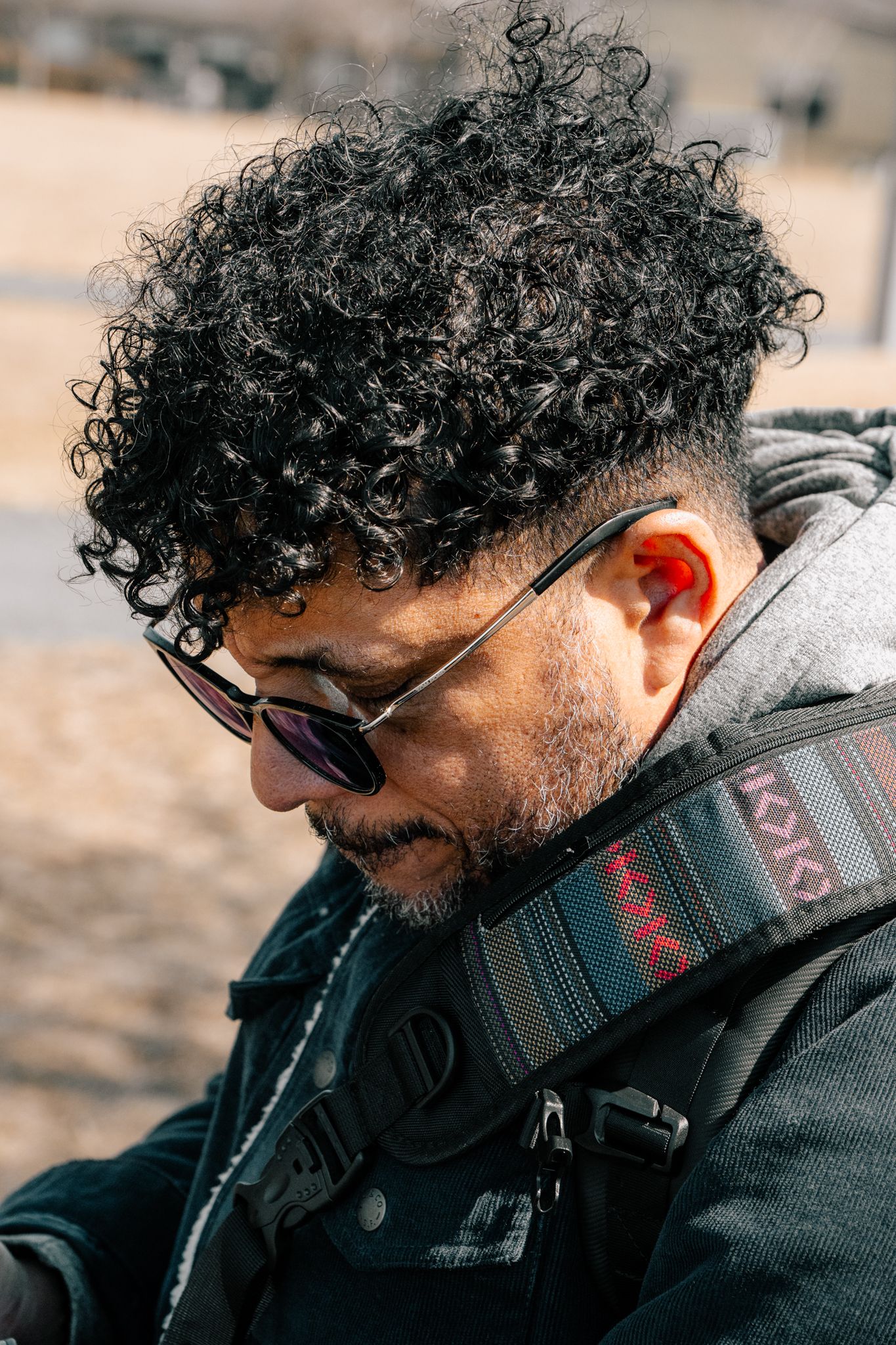 Close-up portrait of curly-haired man with sunglasses and camera strap in sunlight.