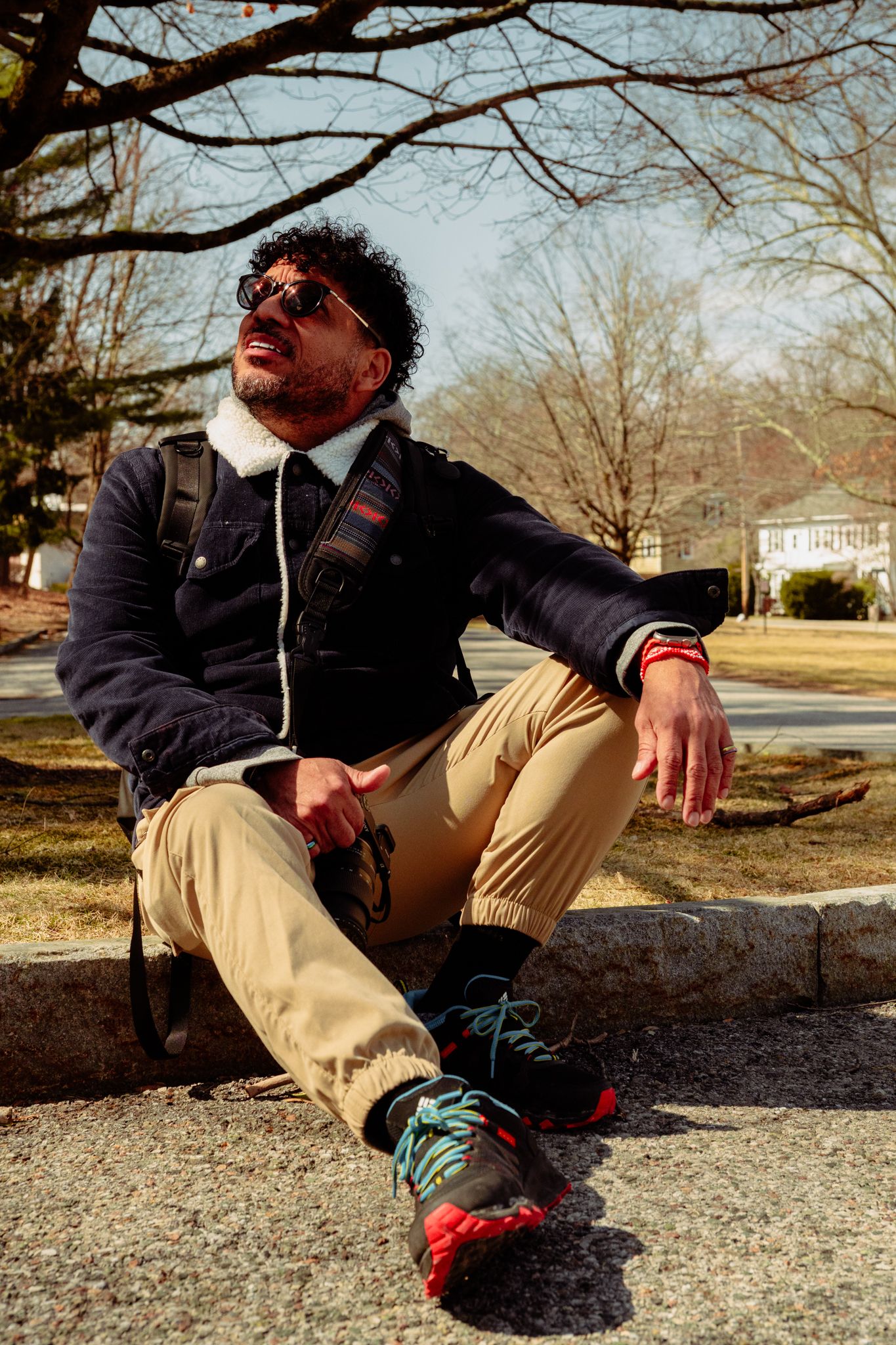 Man in sunglasses and navy jacket relaxing on a park curb, looking up on a bright winter day.