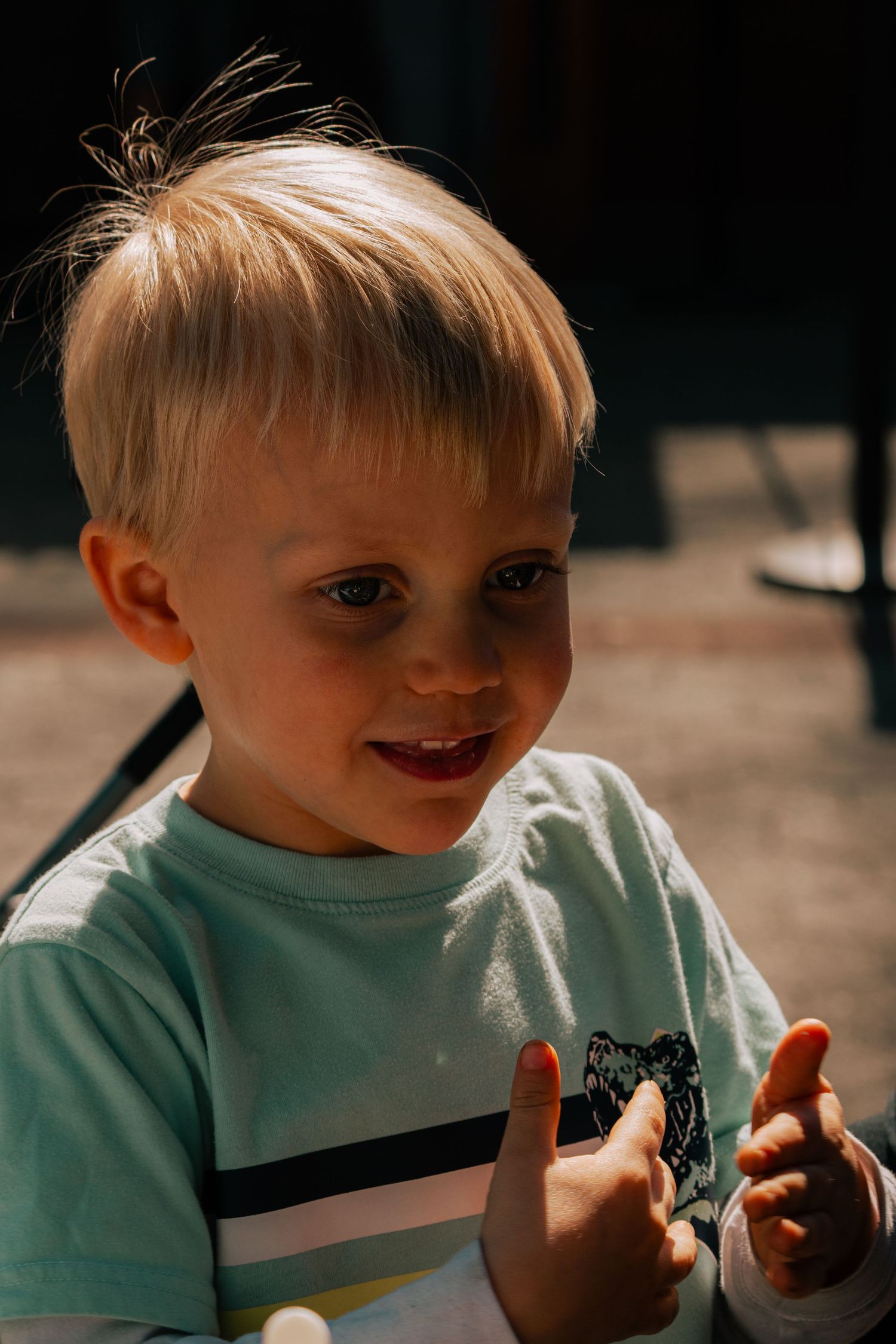 A cheerful blonde toddler boy in a striped light blue shirt, clapping hands and smiling in warm sunlight.