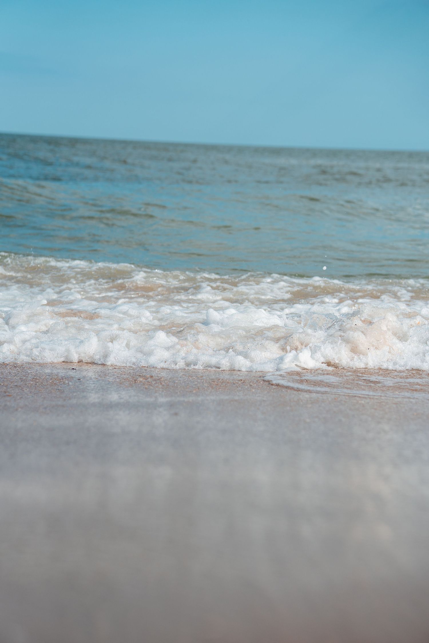 Calm ocean waves with white sea foam washing onto a sandy beach under a clear blue sky.