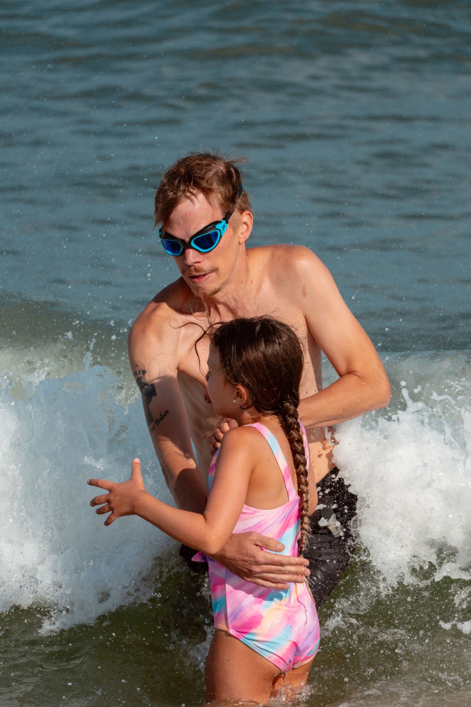 Adult with goggles holding a young girl in a pink swimsuit as ocean waves splash around them.