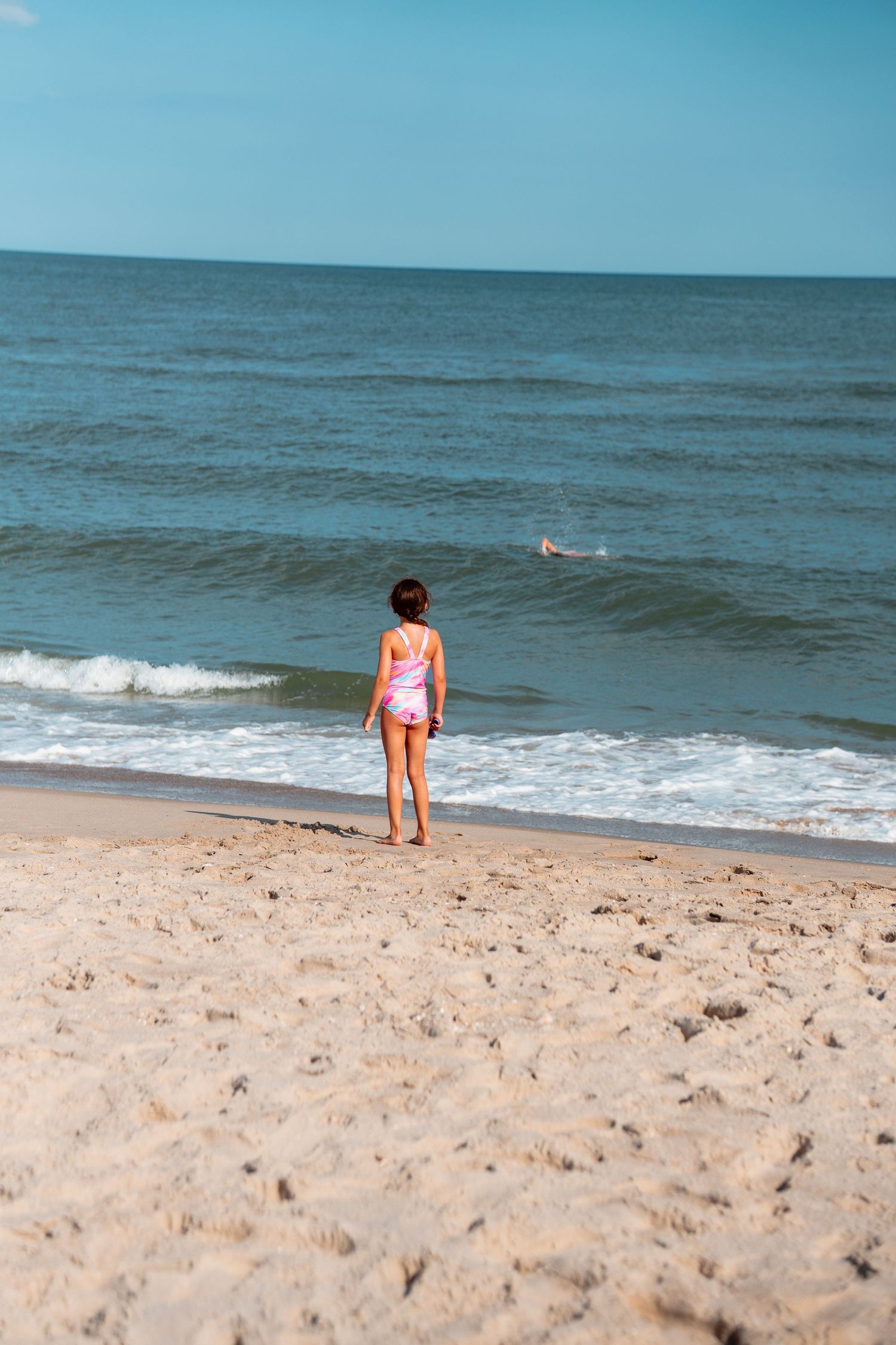 Young child in pink swimsuit standing on sandy beach gazing at ocean waves from shoreline.