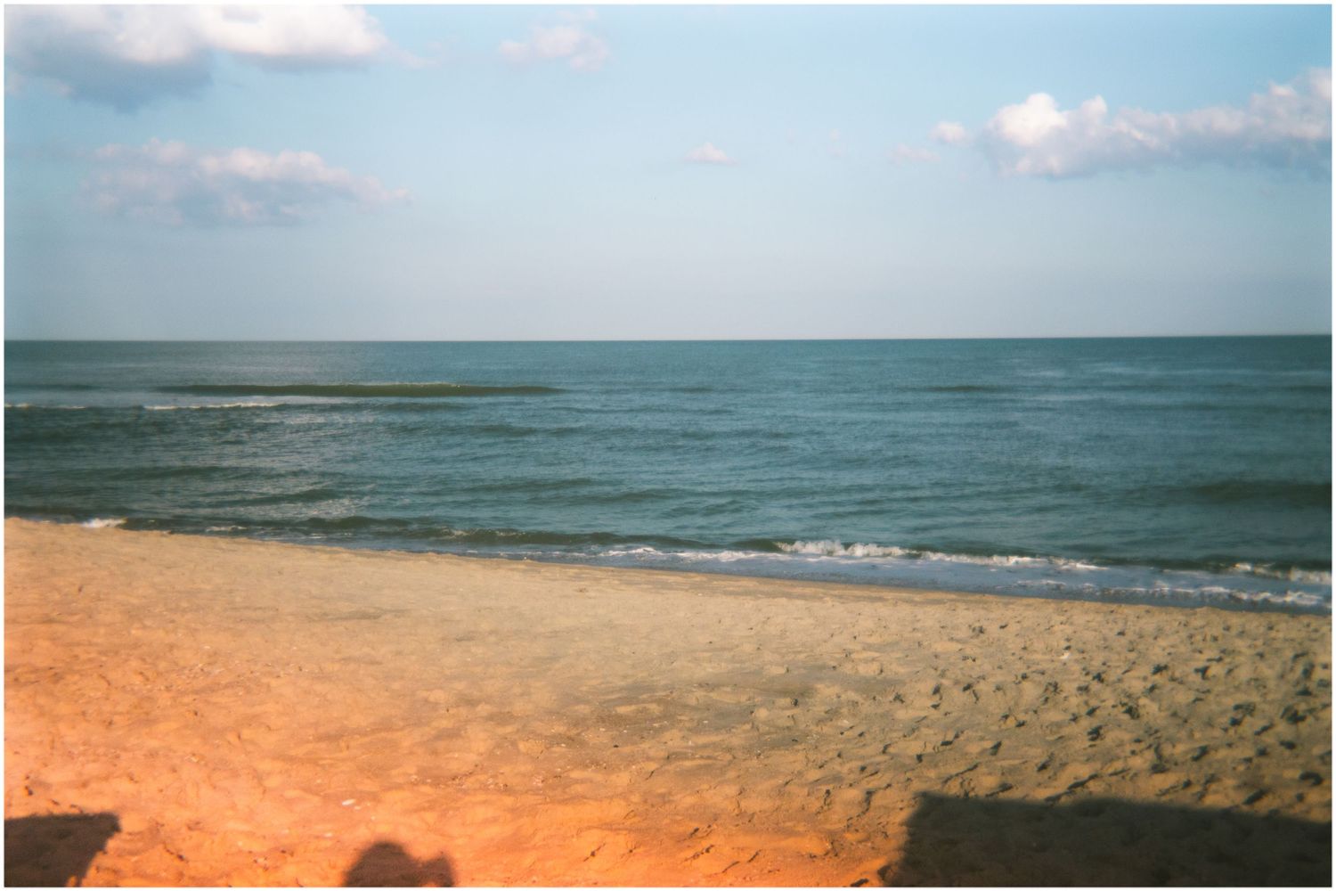 A warm golden sandy beach meets calm ocean waves under a partly cloudy sky at sunset.