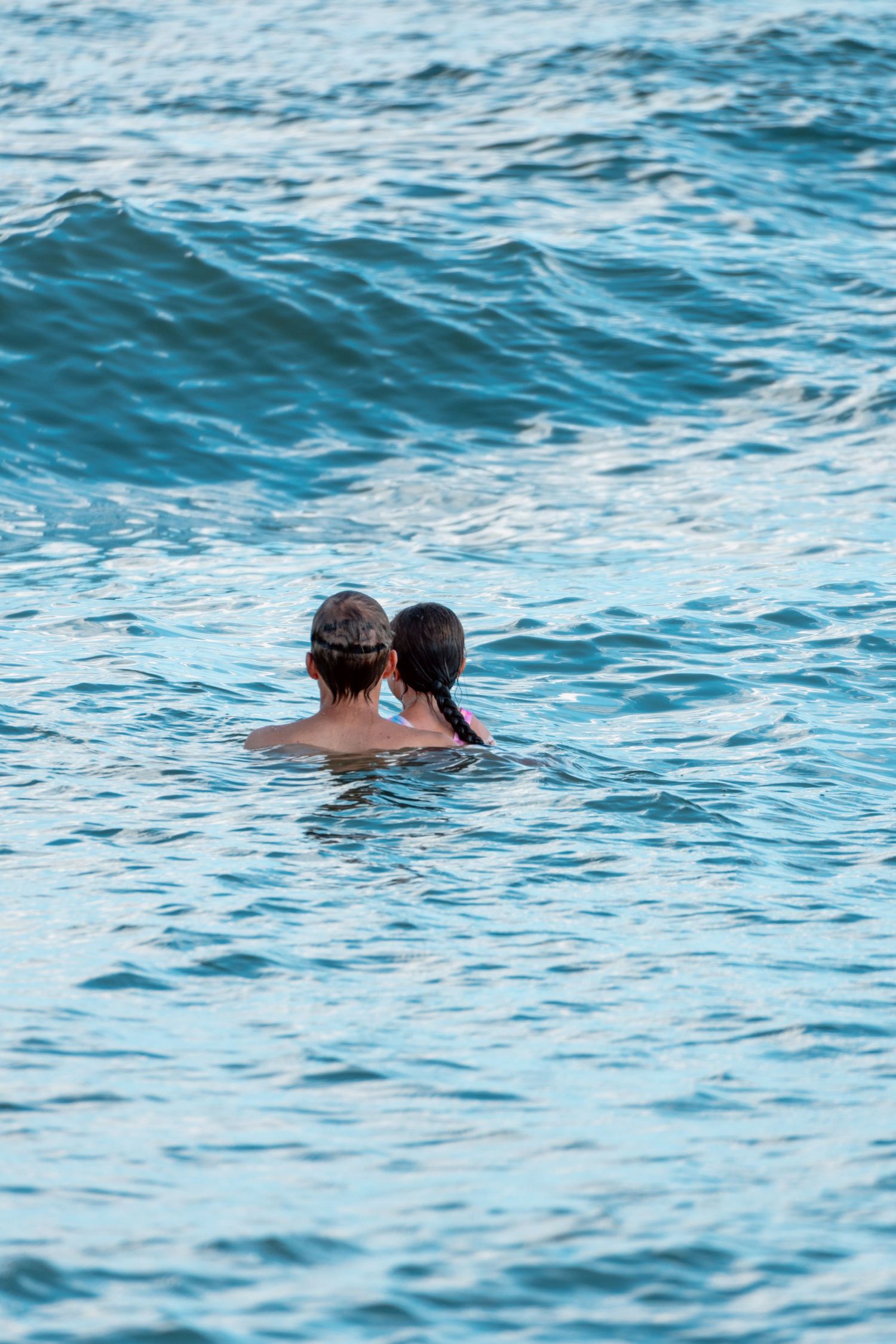 Two people swimming close together in calm blue ocean water from behind.