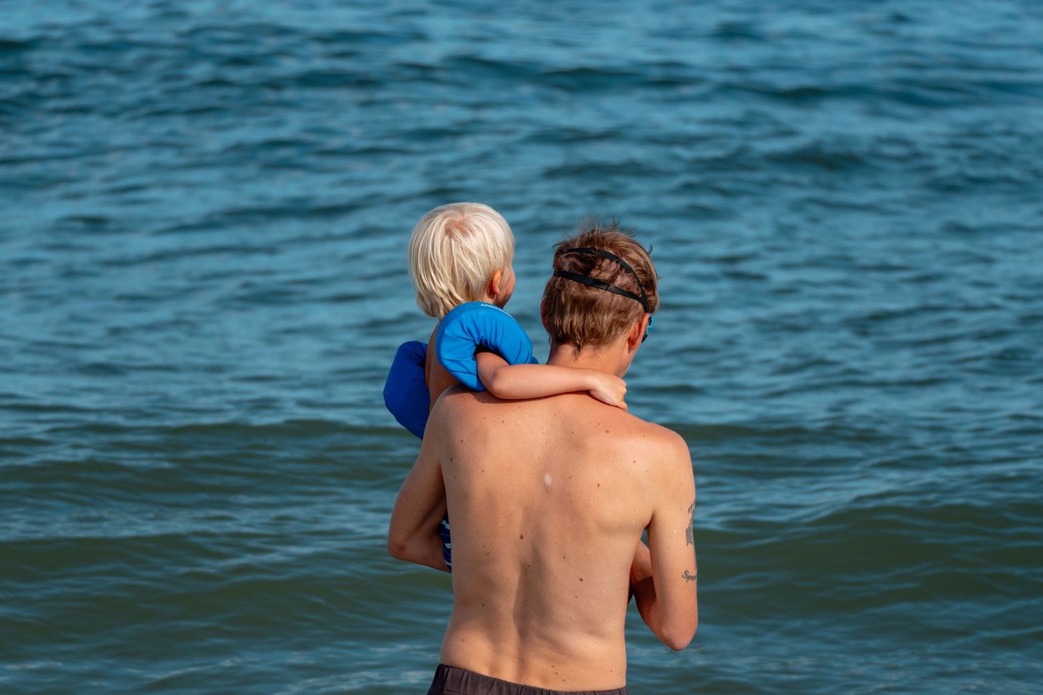 Father carrying young blonde child in blue floaties while standing at ocean edge.
