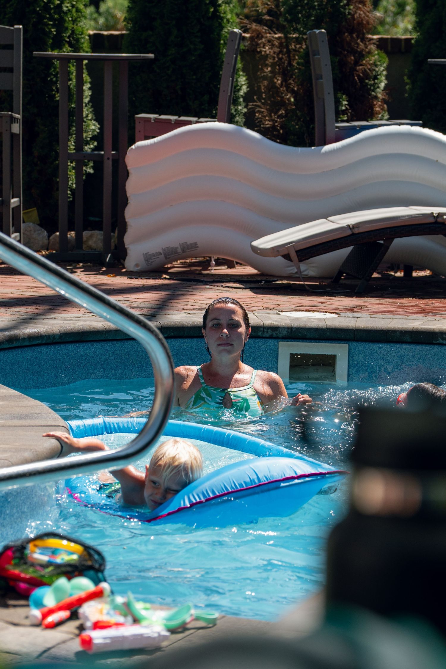 Woman and young child float on blue inflatable raft in backyard swimming pool.