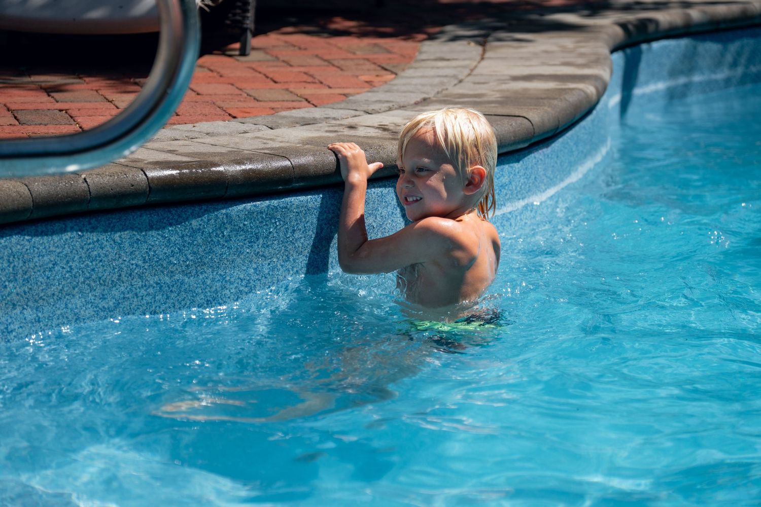 Smiling blonde child holding onto pool edge in a bright sunny backyard swimming pool.