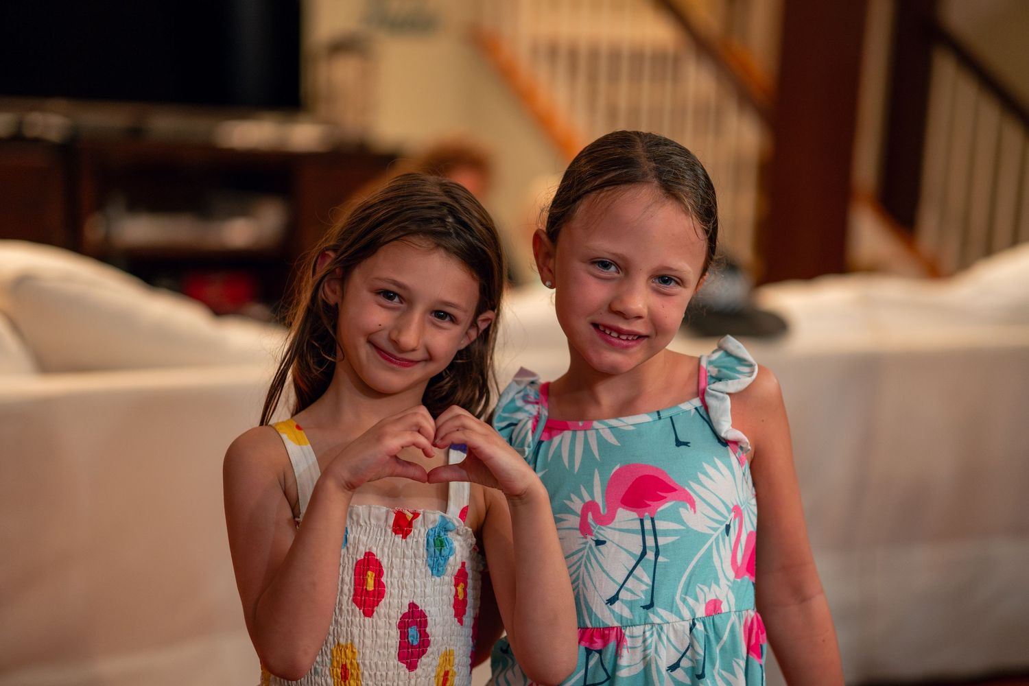 Two smiling young girls making a heart shape with their hands in a cozy living room.
