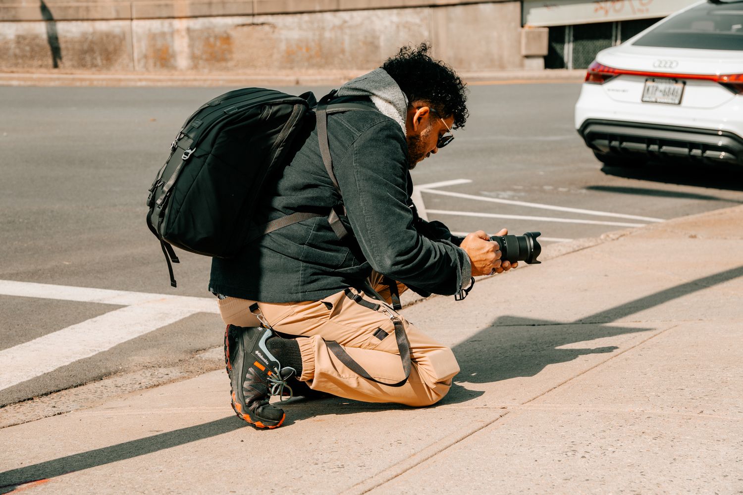 A photographer bending low on a city curb to take a ground-level shot with a professional camera.