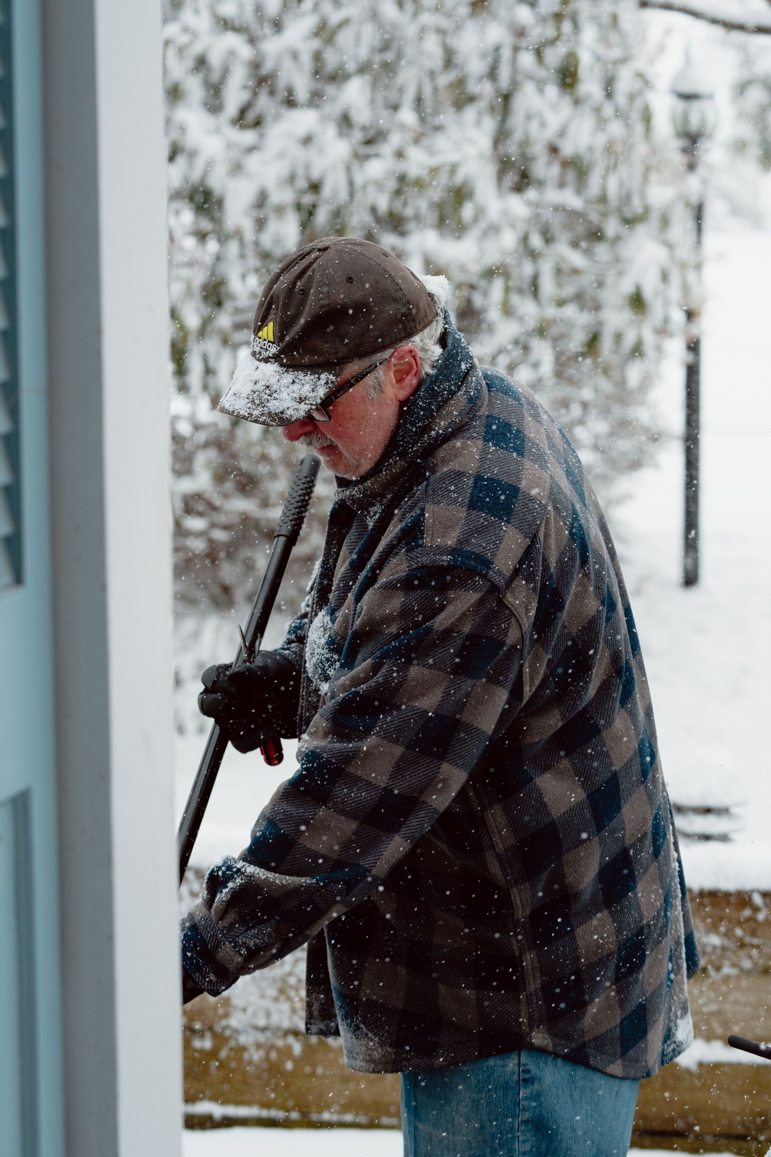Man in plaid jacket shoveling snow off a porch step during a heavy winter snowstorm.