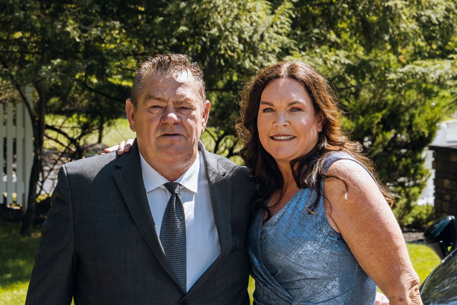 A smiling couple dressed in formal attire poses outdoors at a sunny garden event.