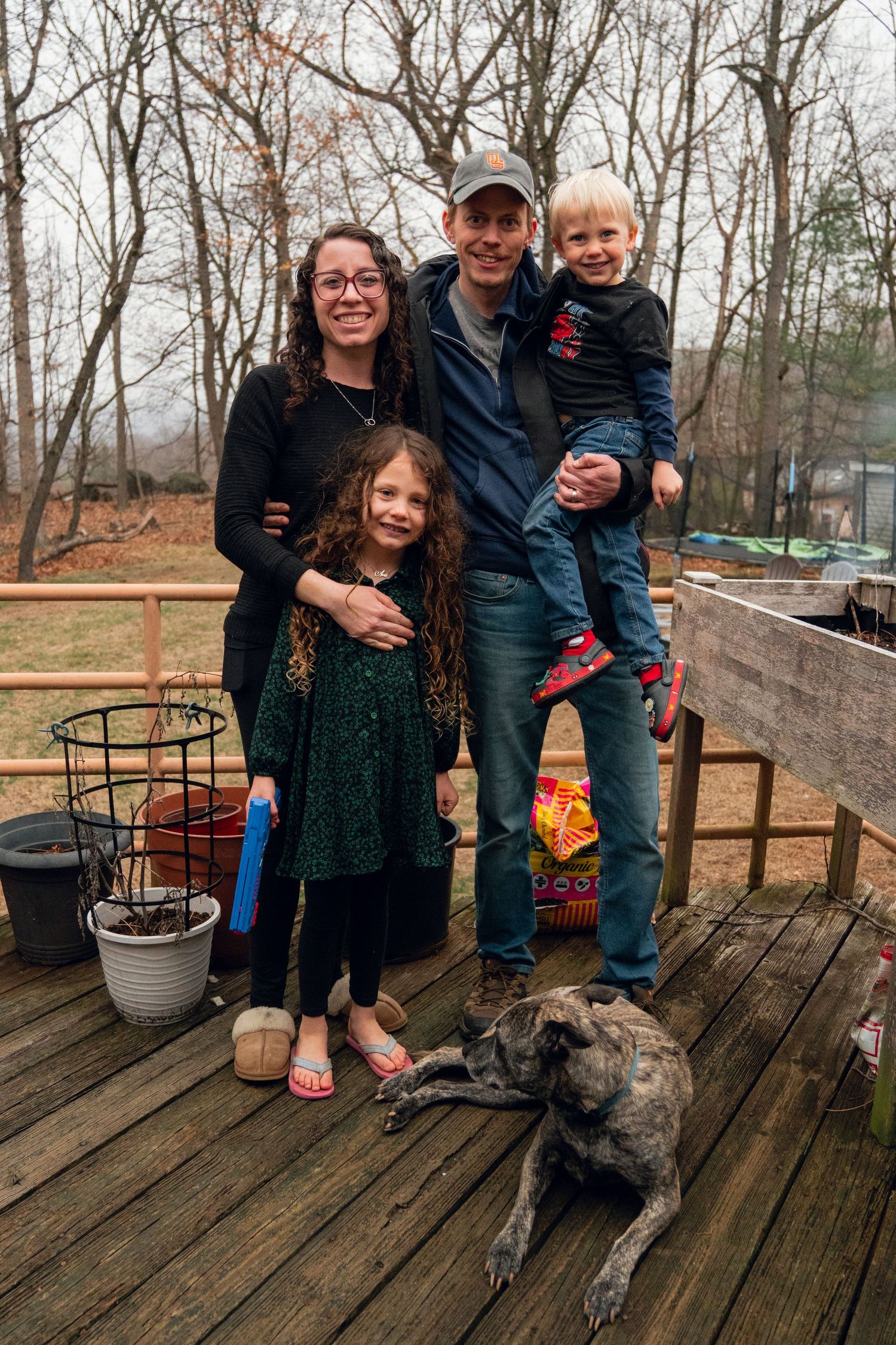 Family of four with a dog posing on a wooden deck surrounded by bare trees on a cloudy day.