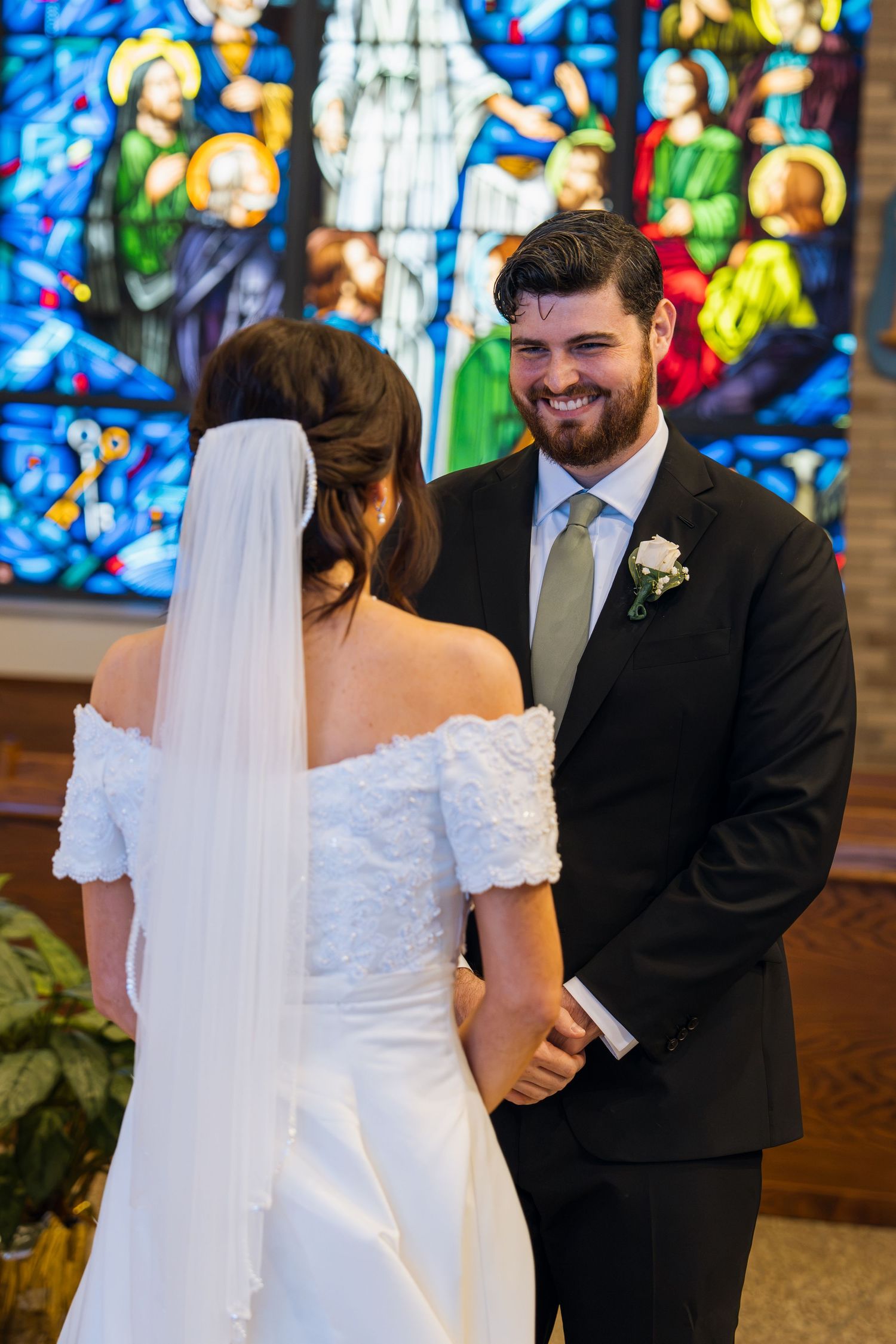 Smiling groom holds bride's hands at church altar during wedding ceremony with stained glass backdrop.