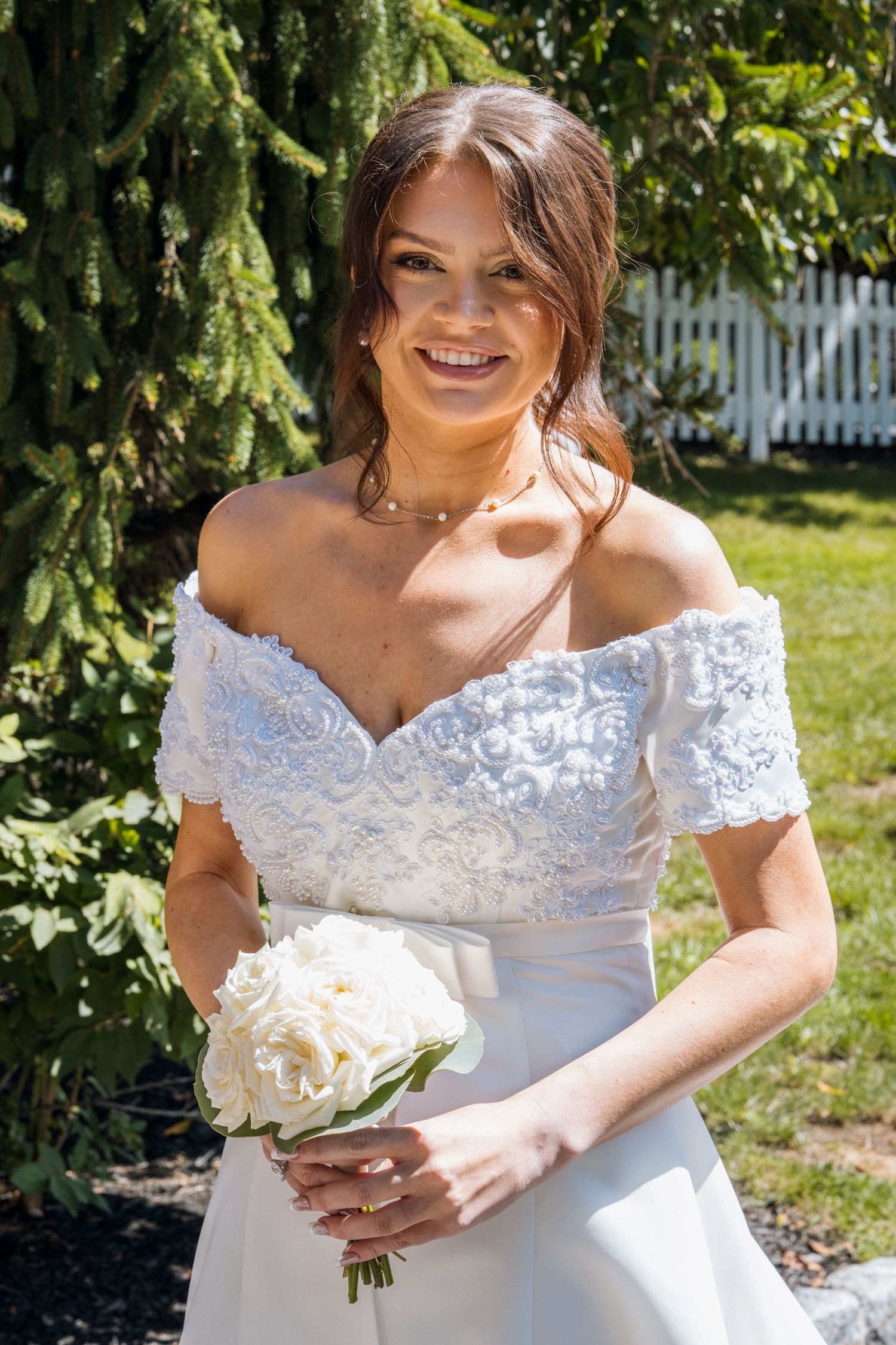 Smiling bride in off-shoulder lace wedding dress holding white peony bouquet outdoors near evergreen tree.