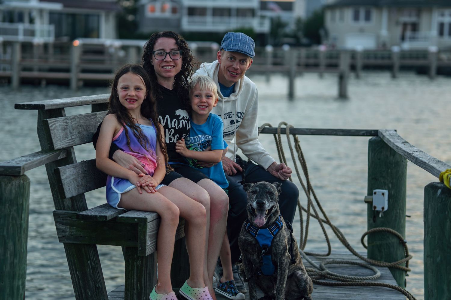 Family of four with a dog sitting on a wooden dock by the water at dusk, smiling together.