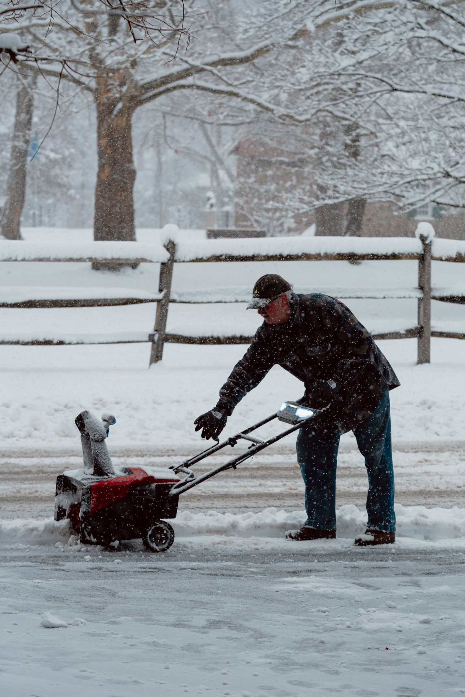 A man operating a red snowblower to clear a snow-covered driveway during a winter storm.
