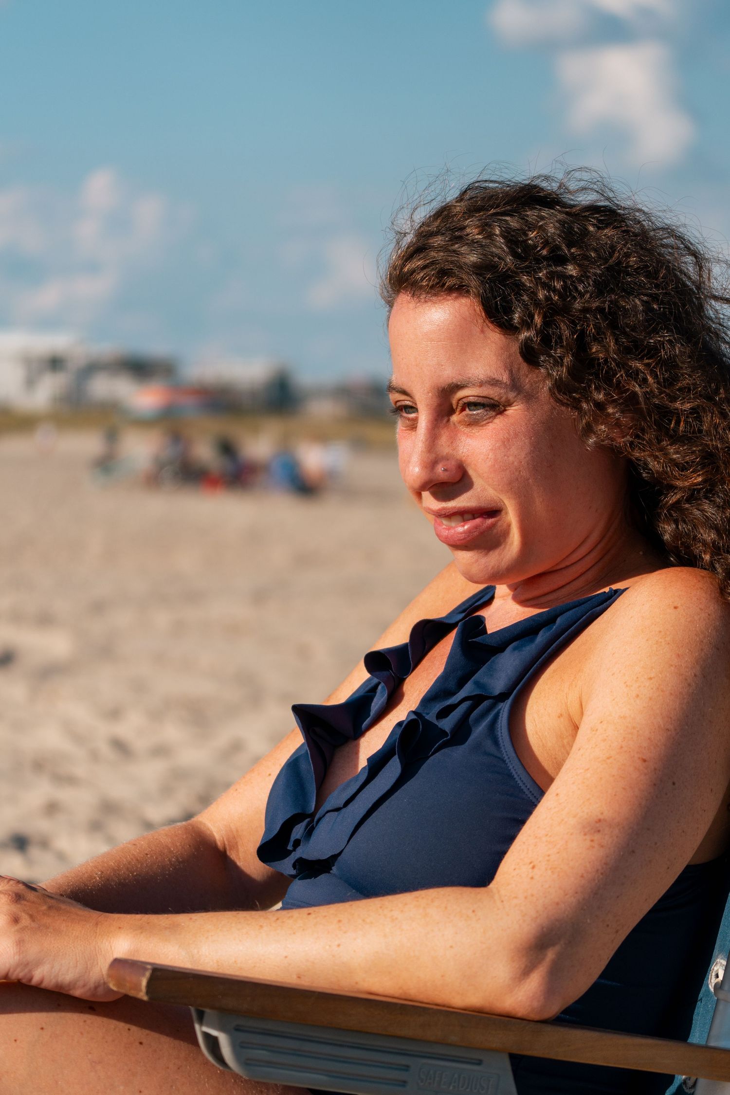 A woman with curly hair in a navy swimsuit relaxes on a beach chair outdoors.