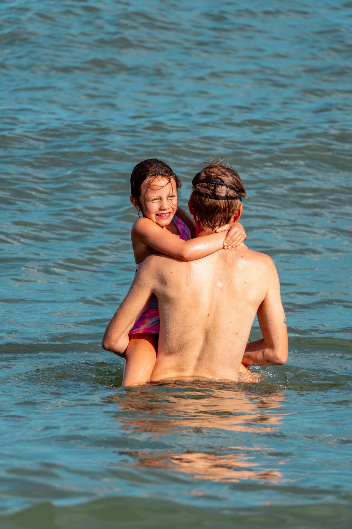 Smiling young girl clinging to an adults back while playing in sparkling blue ocean water.