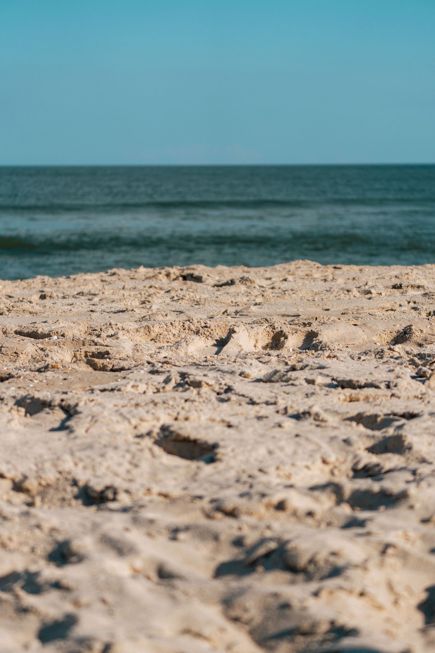 Close-up of sandy beach with textured sand in the foreground and calm ocean waves under a clear blue sky.