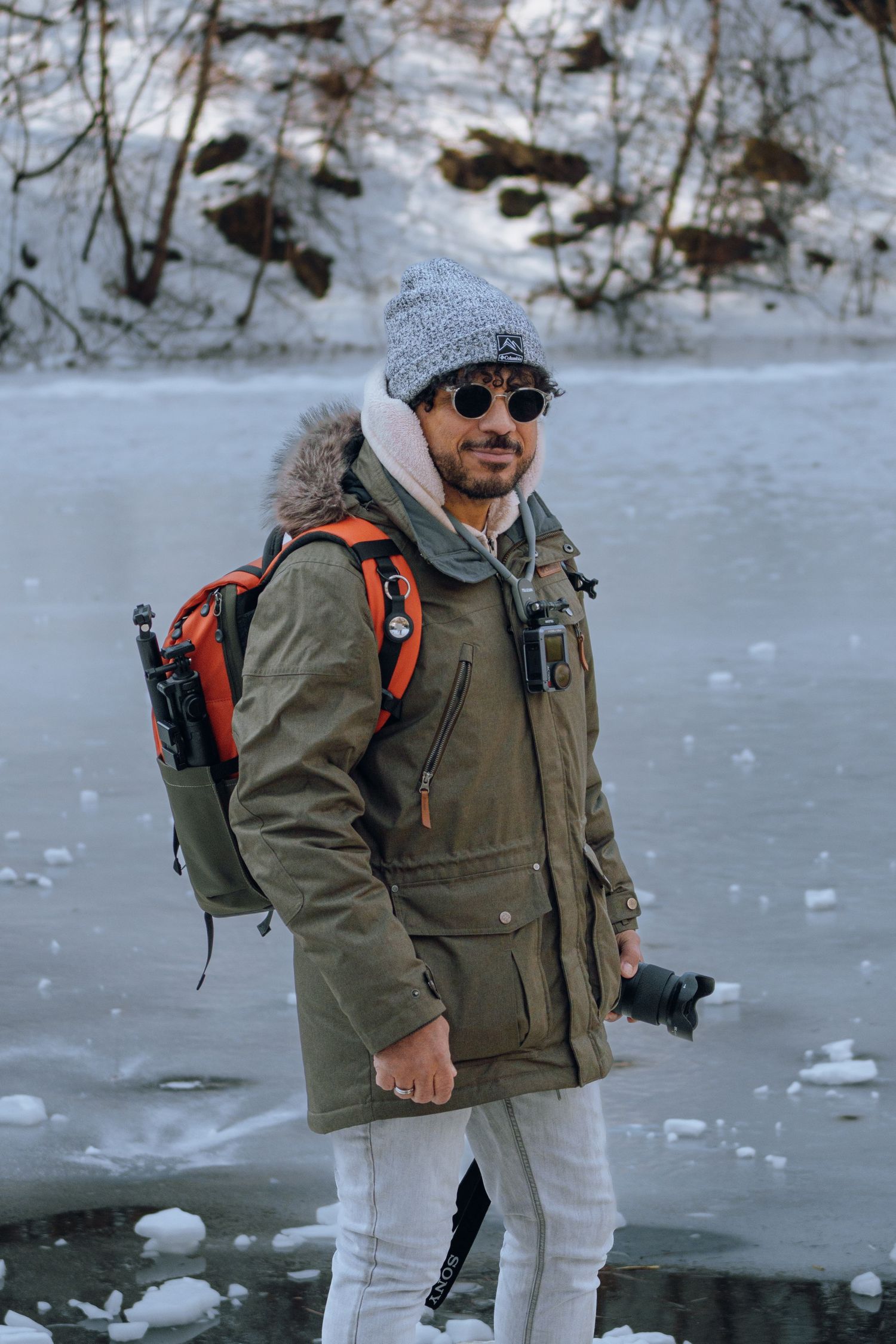 Male photographer in winter parka with orange backpack poses near frozen snowy lake outdoors.
