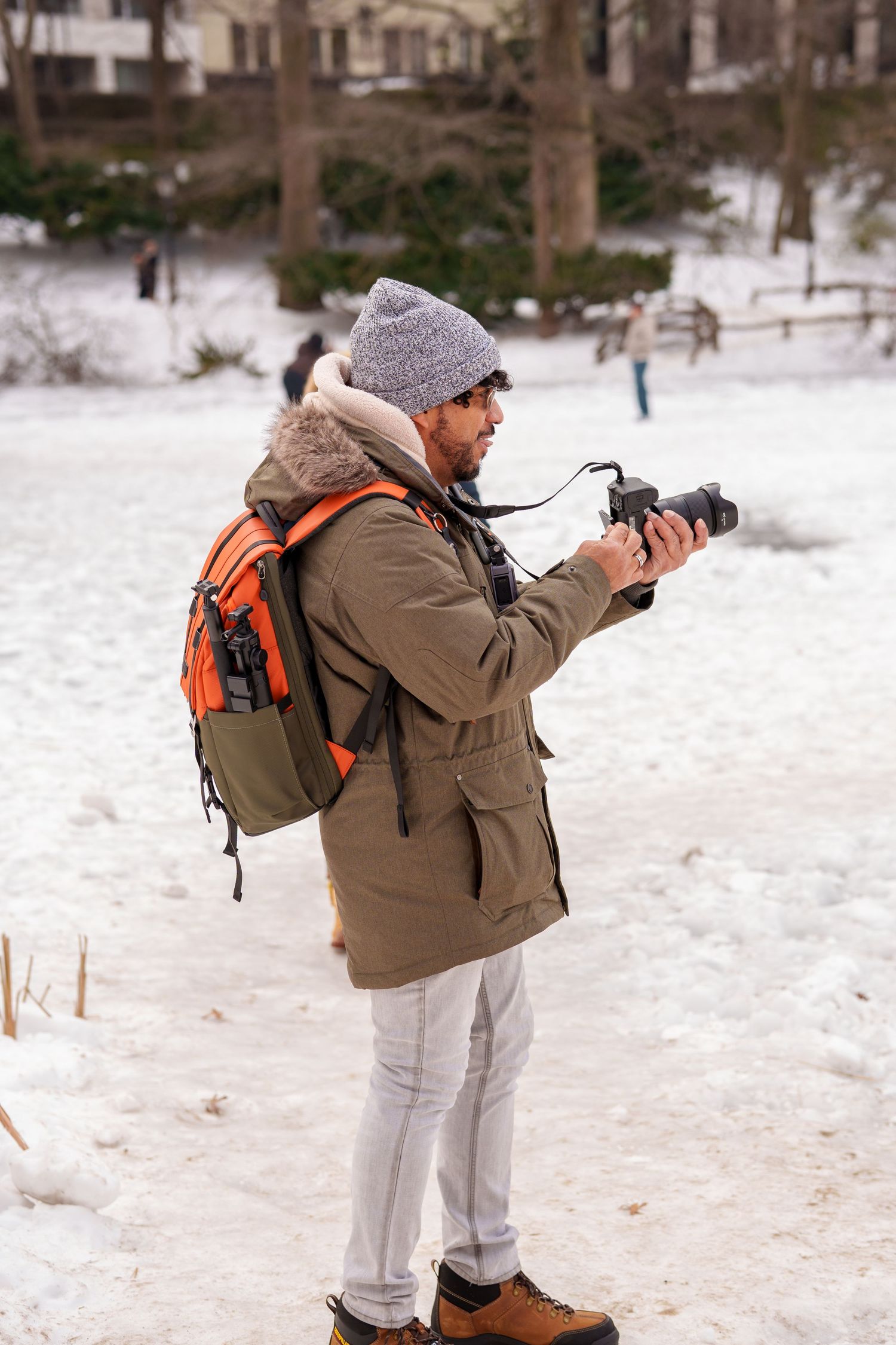 Bearded photographer in olive parka with orange backpack shooting photos in a snowy park.