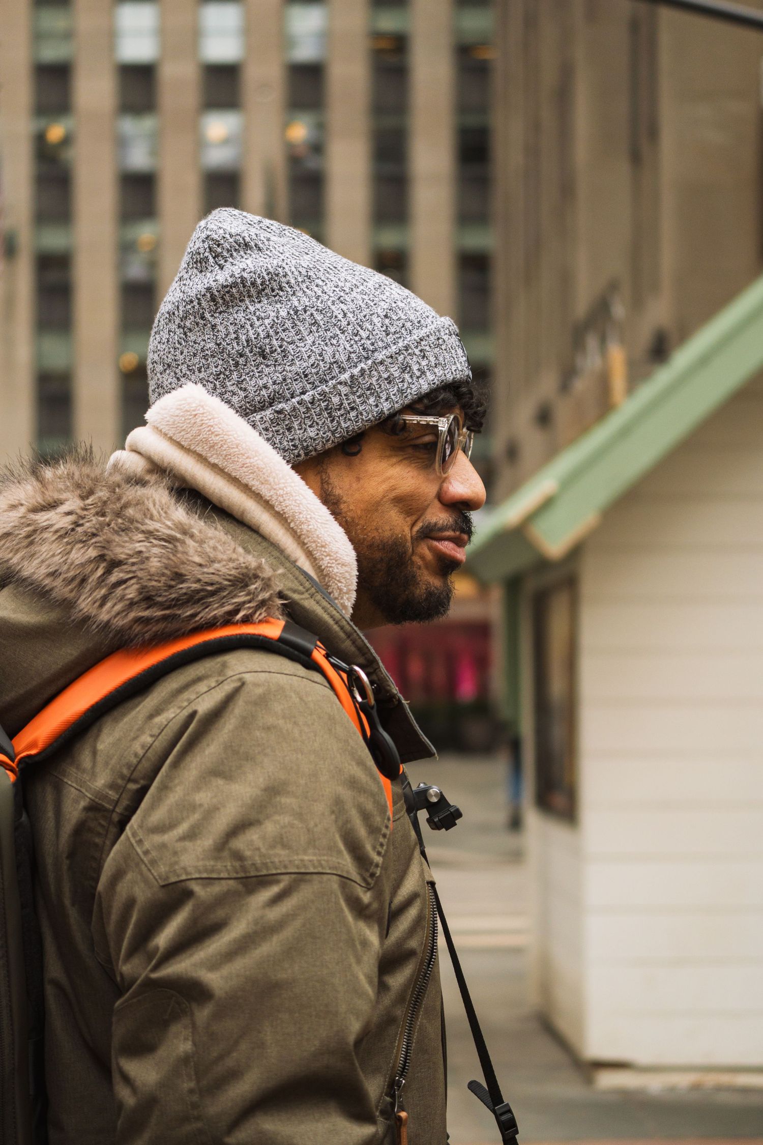 Bearded man in gray beanie hat and olive parka with orange backpack straps in urban city setting.