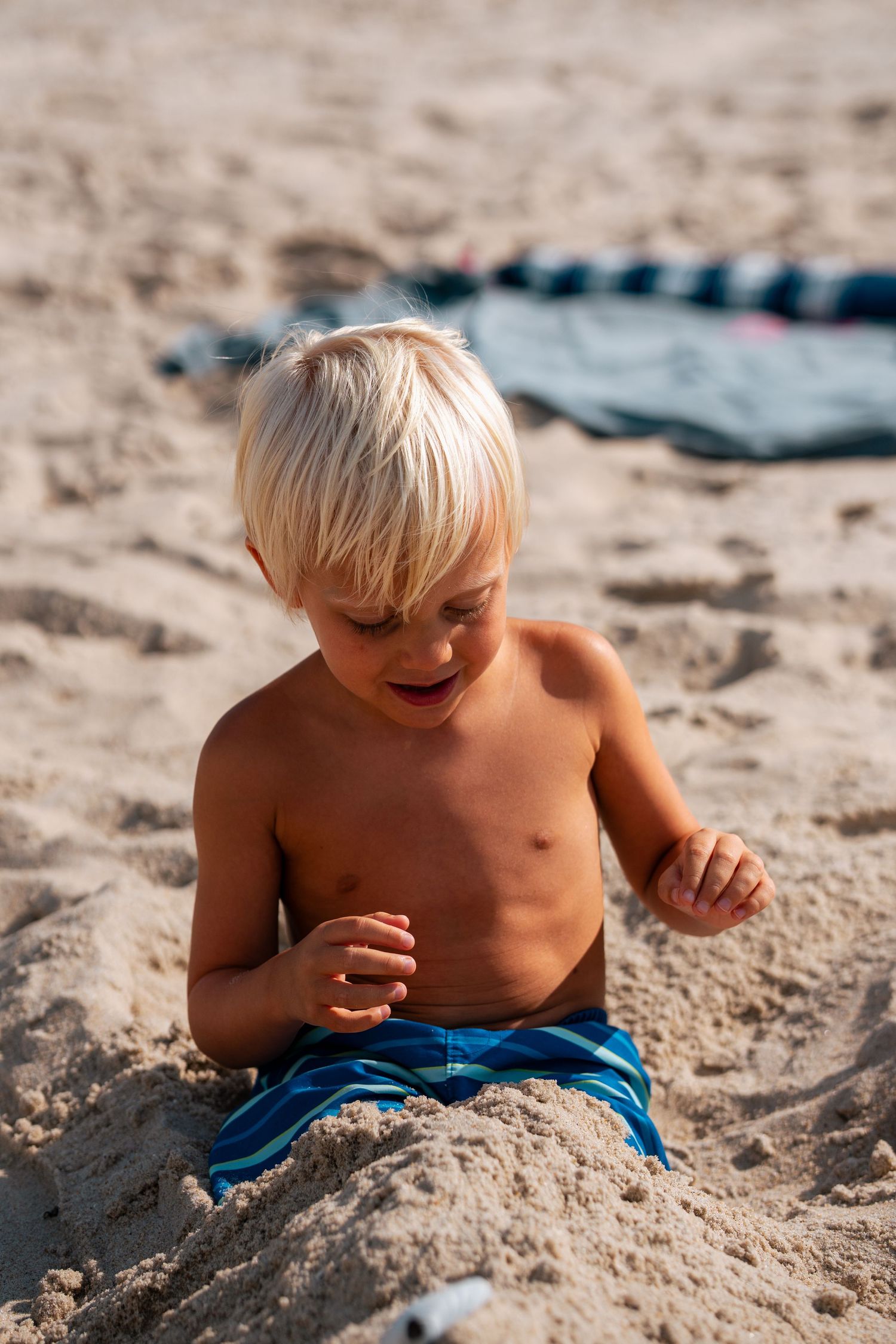 Happy blonde toddler boy in blue swim trunks playing in sand at the beach.