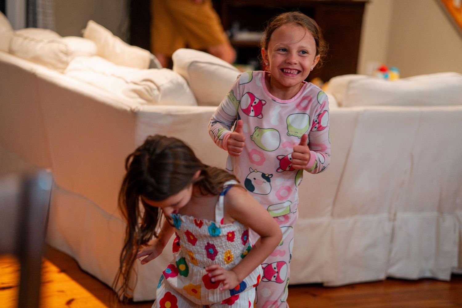 Two girls in pajamas laugh and play together in a cozy living room at nighttime.
