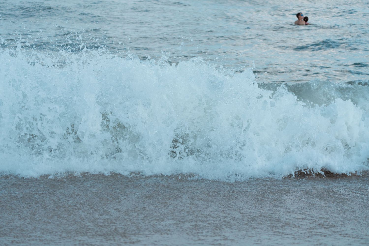 A large breaking ocean wave with two swimmers visible in the background.