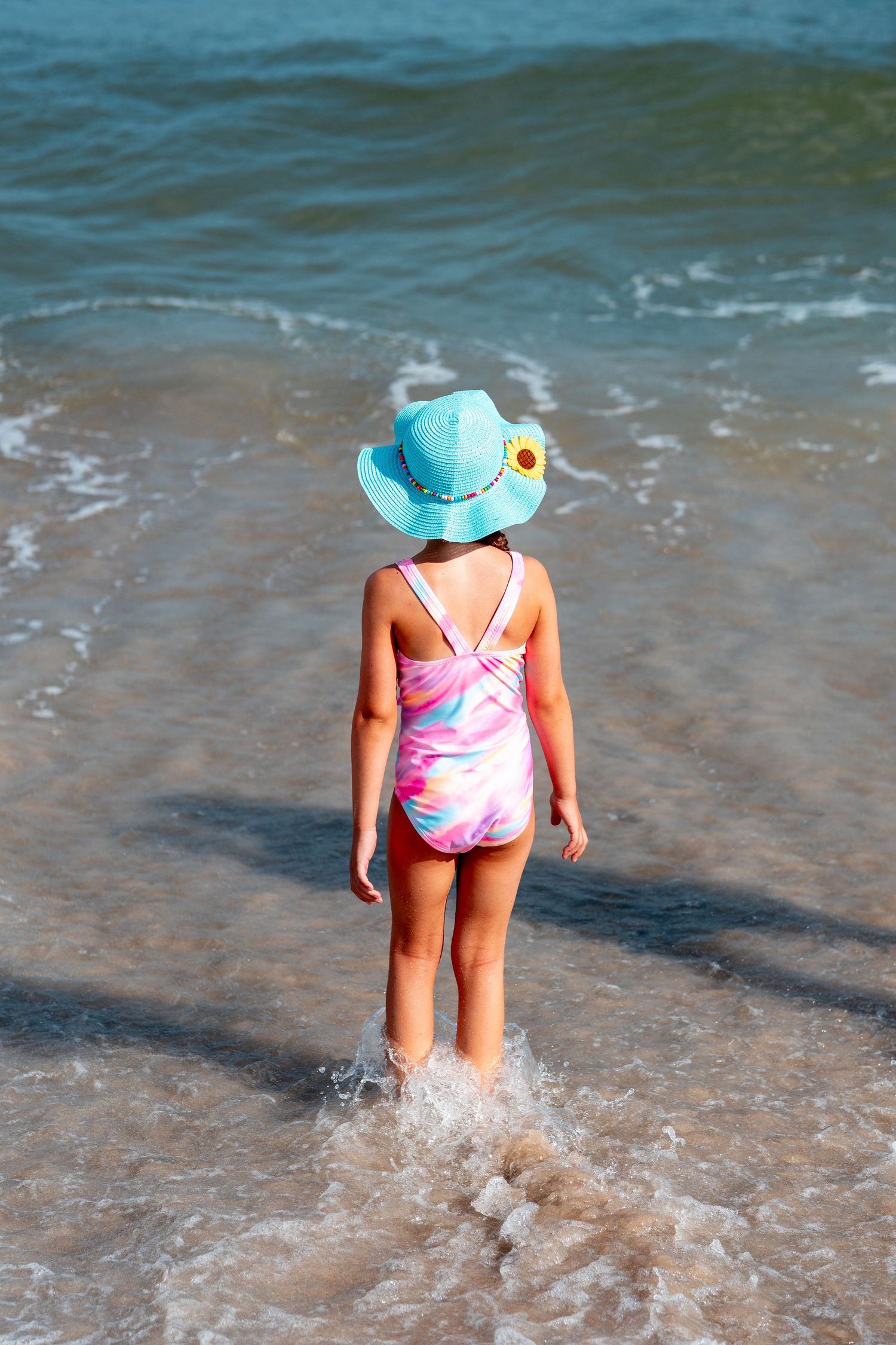 Young girl in blue bucket hat and colorful swimsuit standing in shallow ocean waves.