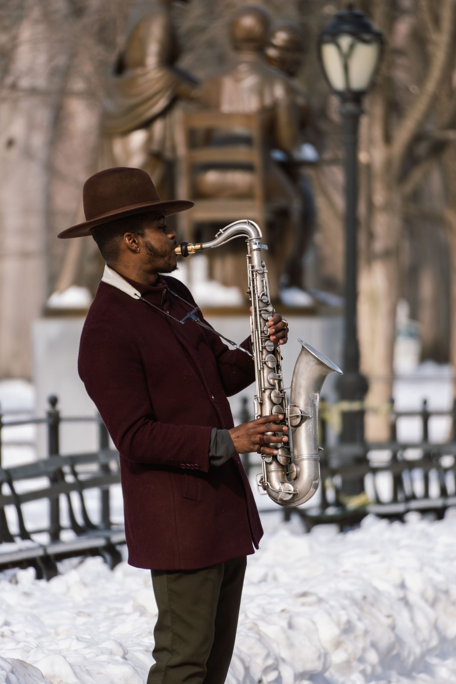 A saxophonist in a burgundy blazer performs outdoors in a snowy park with a bronze statue behind him.