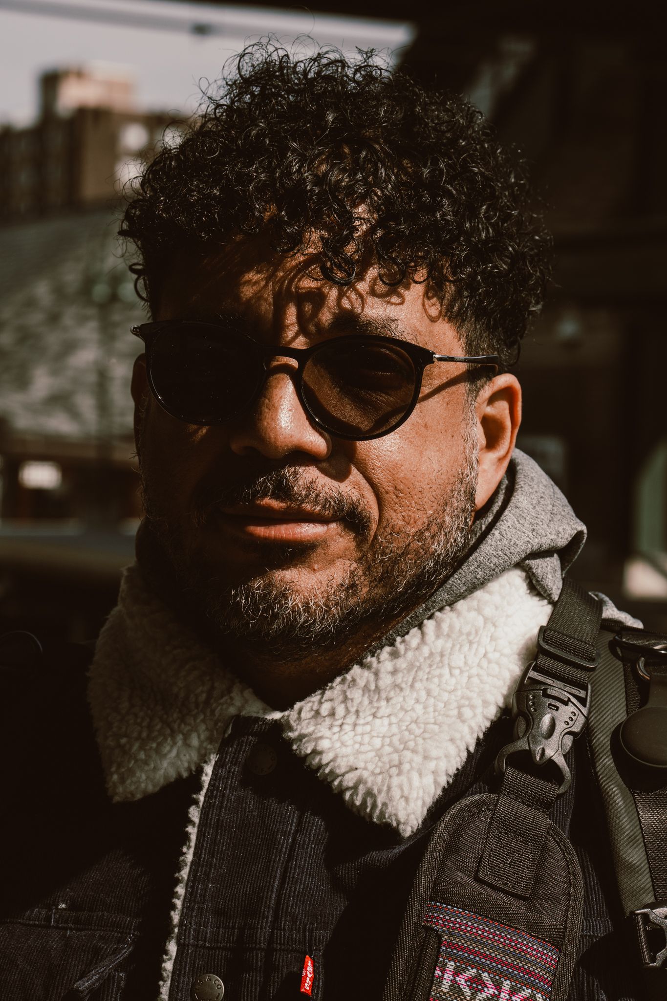 Close-up portrait of a bearded man with curly hair wearing sunglasses and a sherpa-lined denim jacket outdoors.