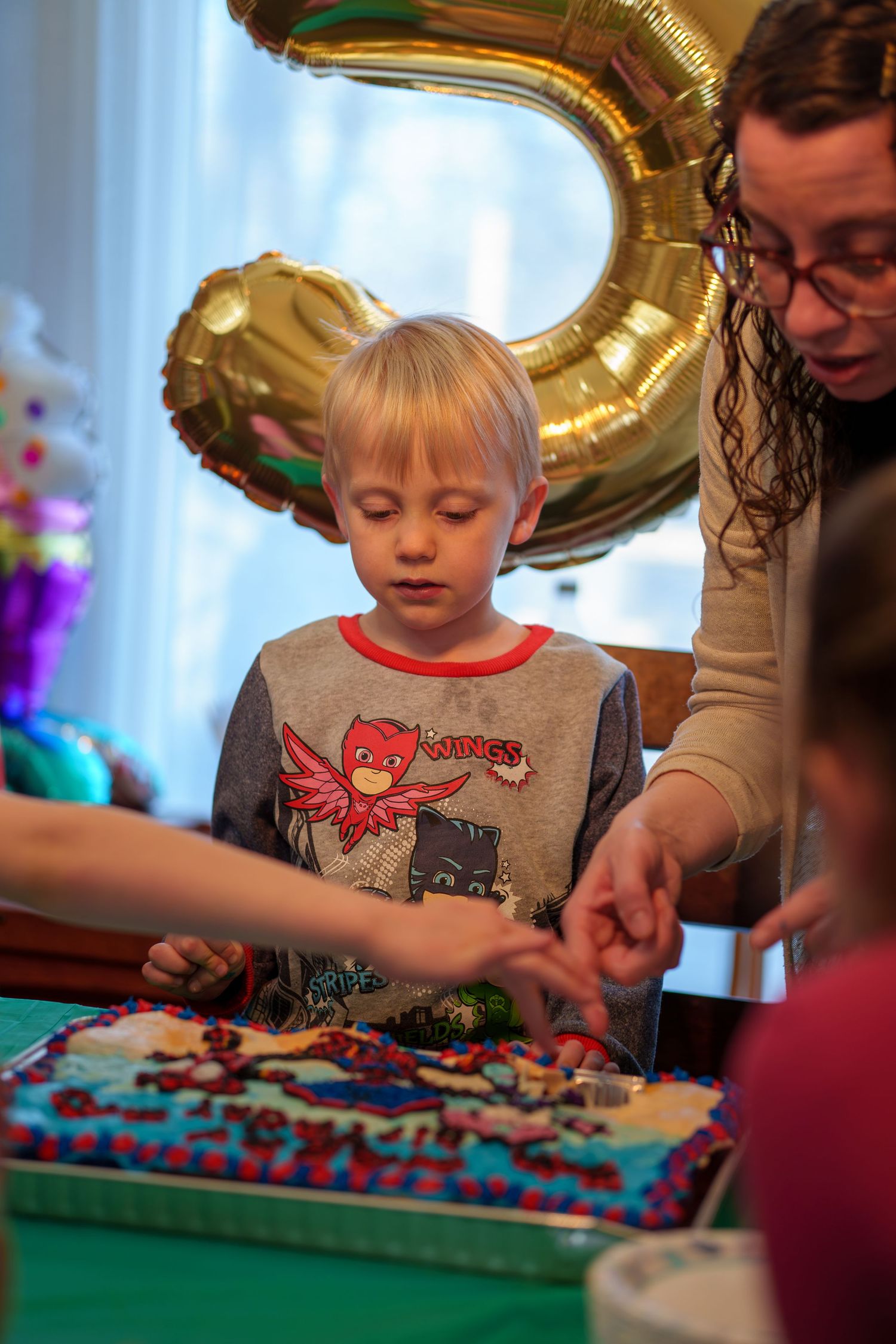 Young boy at 3rd birthday party with decorated cake, gold number balloon, and adult helping celebrate.