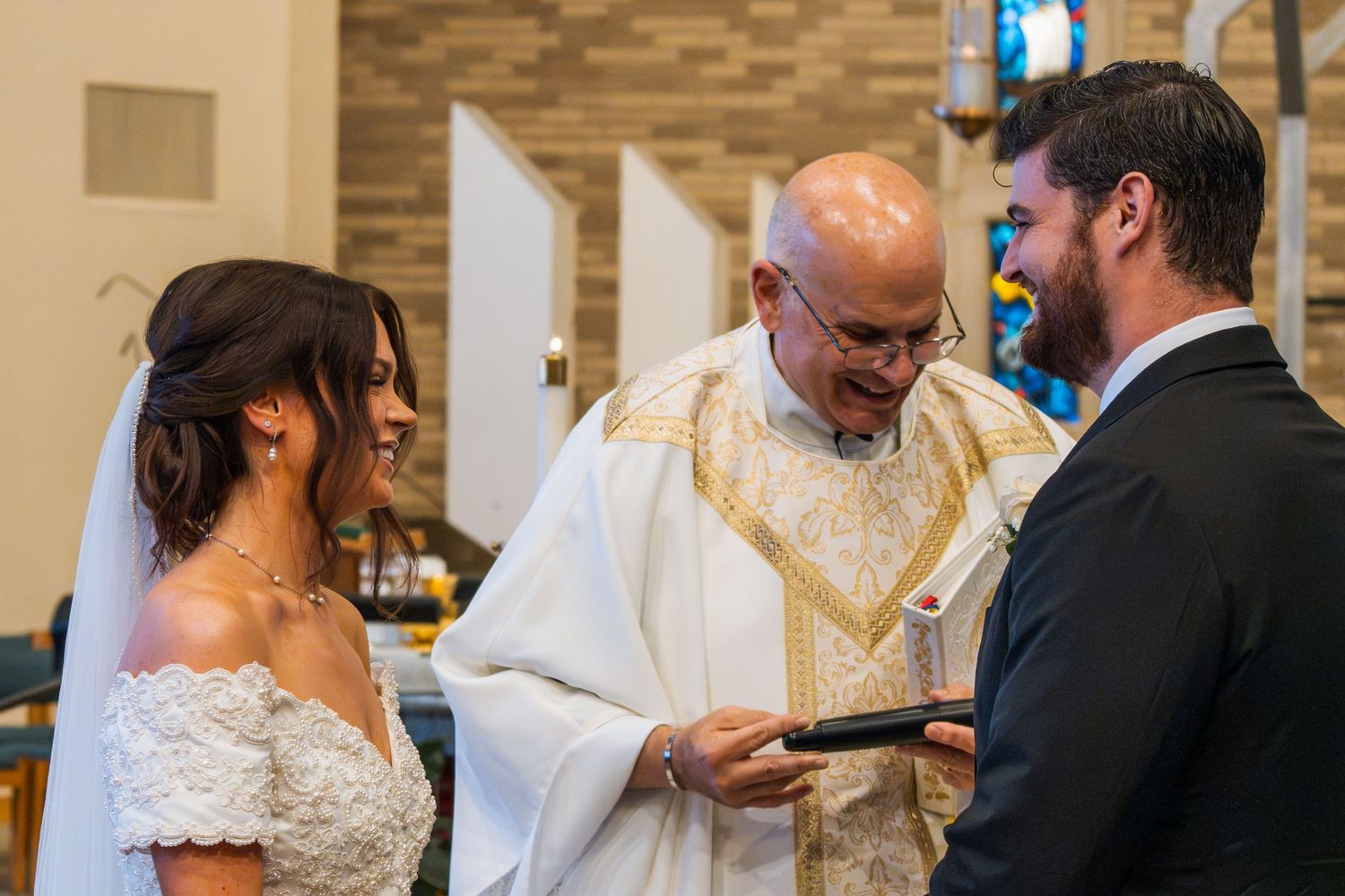 Bride and groom exchanging vows with smiling priest during church wedding ceremony.