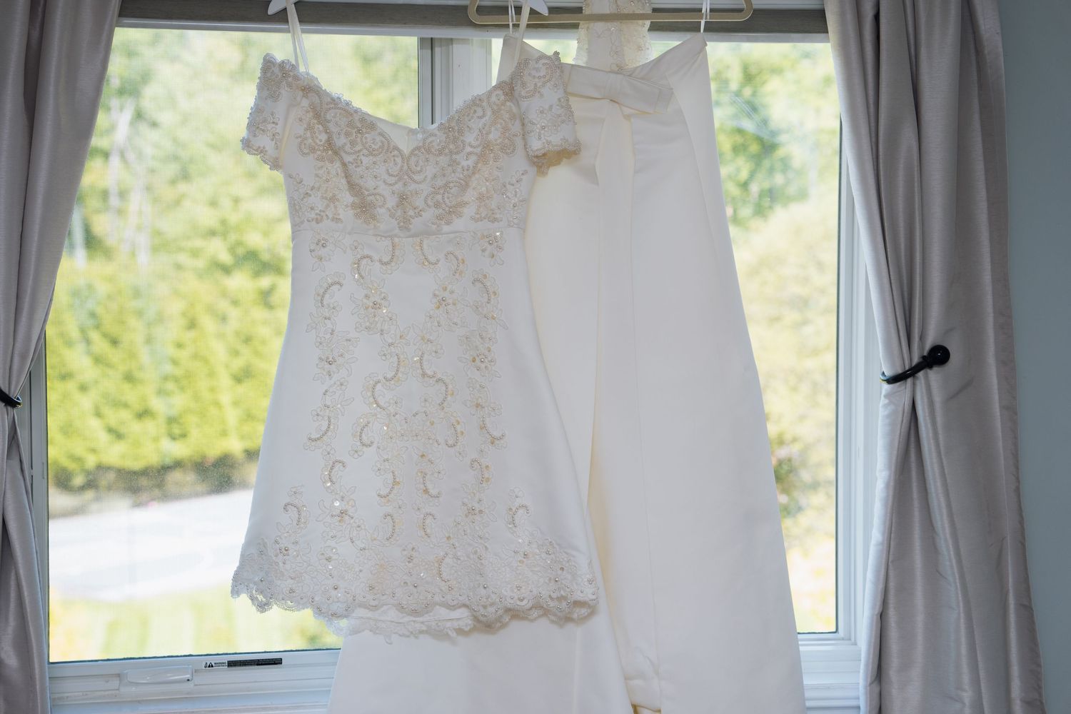 Two elegant white wedding dresses hanging in a sunlit window with sheer curtains and green trees outside.