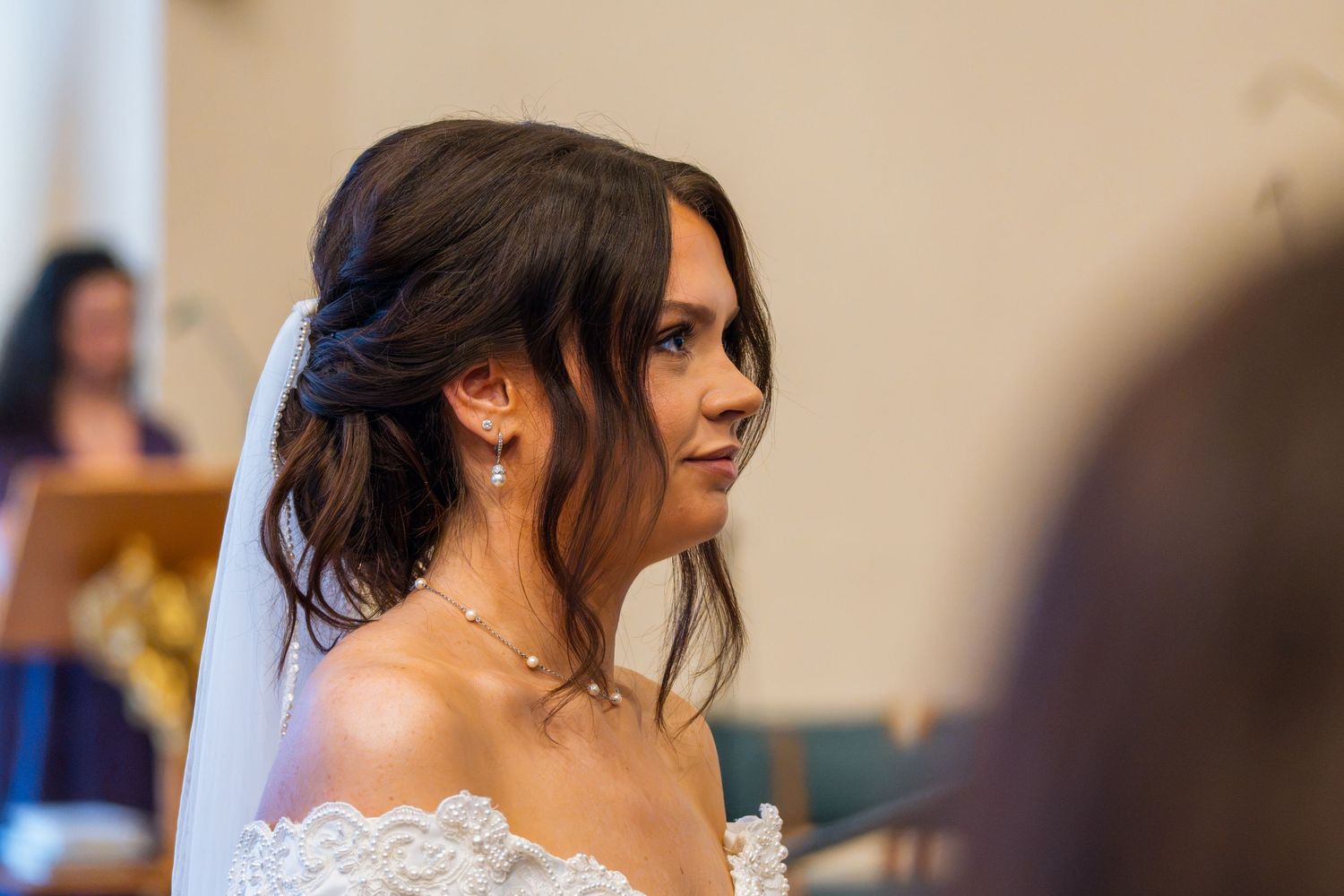 Bride in lace off-shoulder gown with veil and pearl jewelry gazes forward during wedding ceremony.