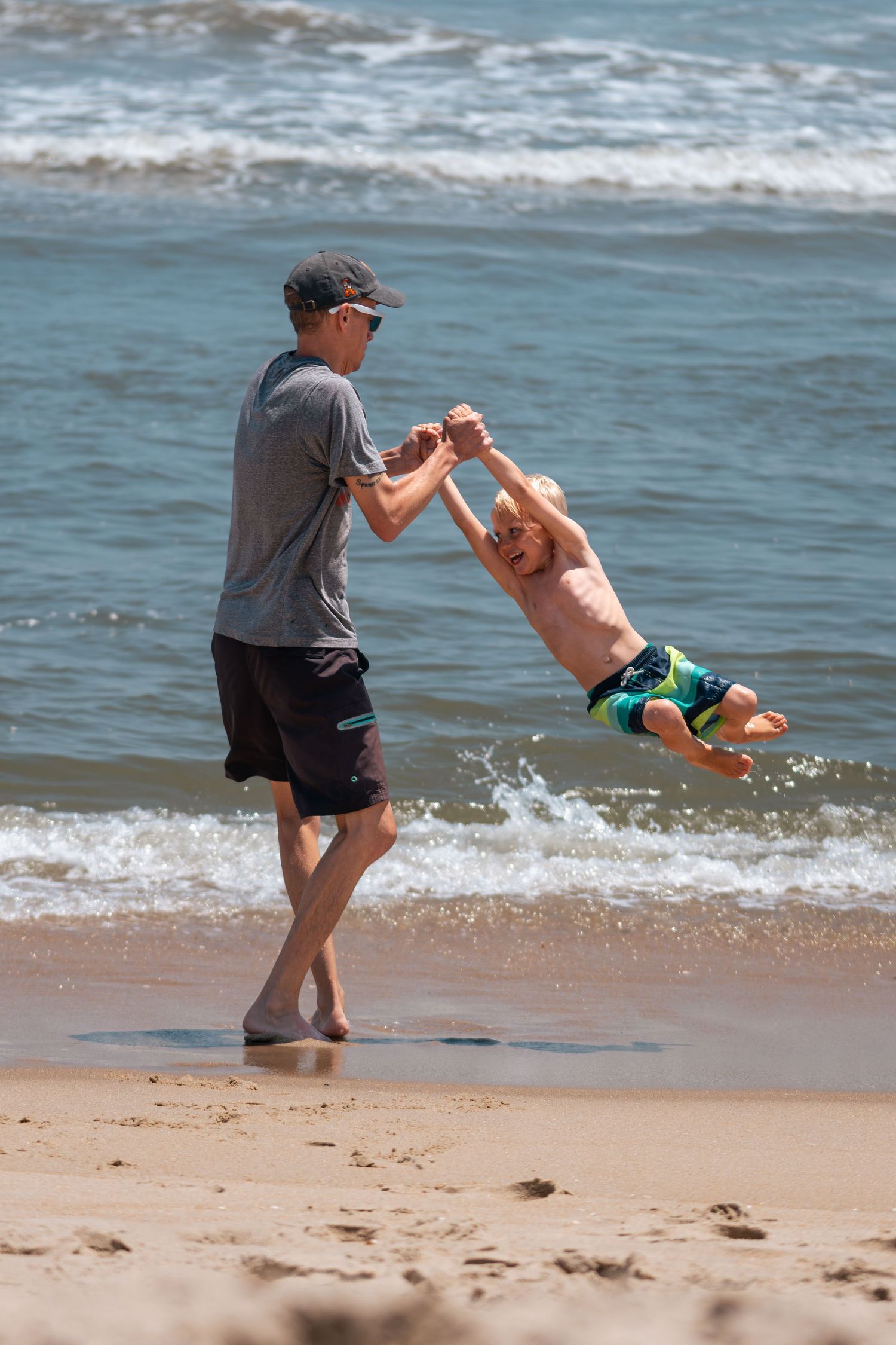 Adult swinging a laughing child by the hands on a sunny beach with ocean waves in the background.