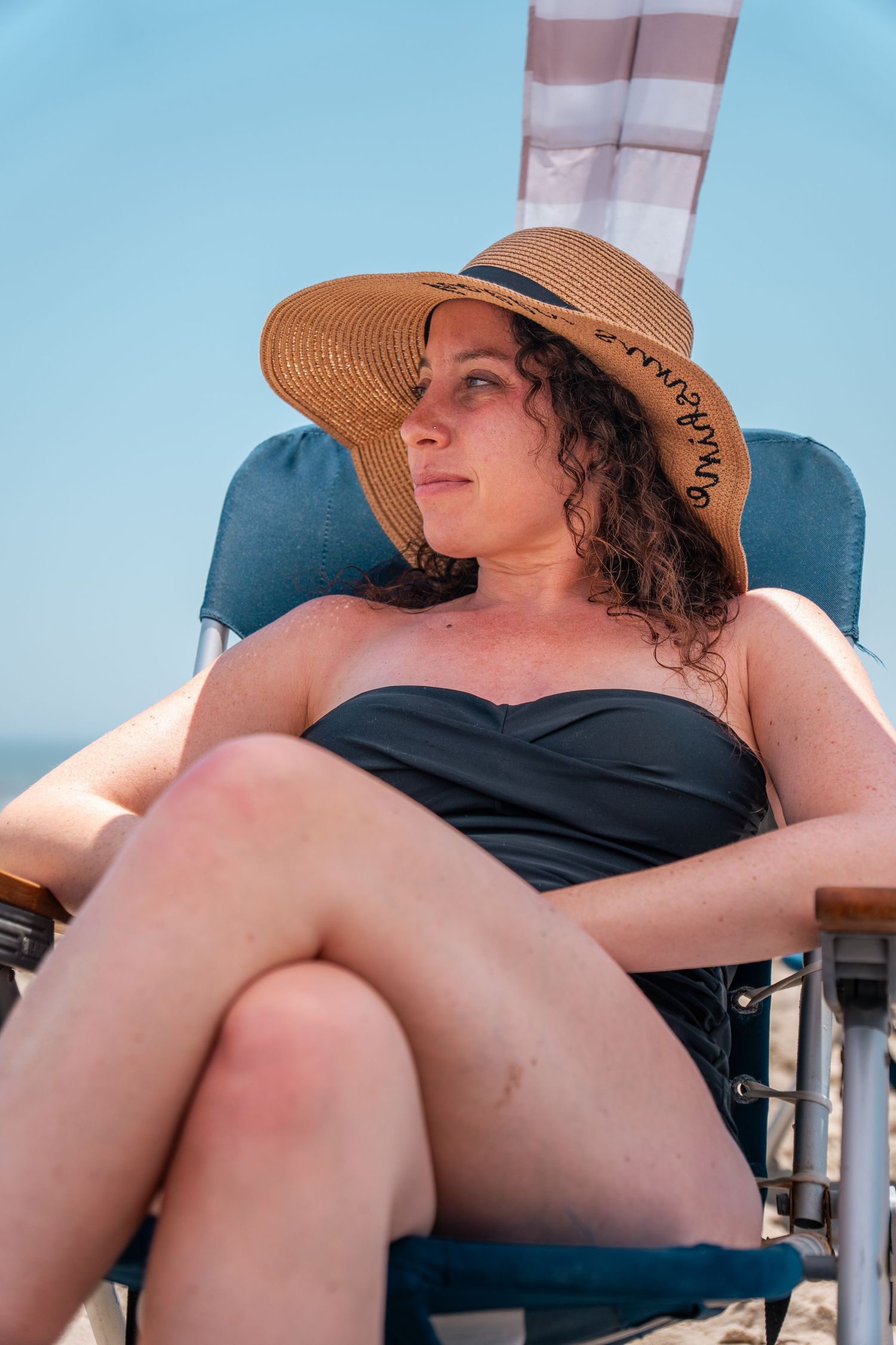 Woman in black swimsuit and straw sun hat relaxing on beach chair against blue sky.