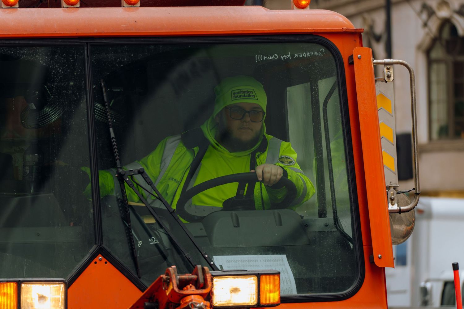 Smiling worker in yellow safety jacket driving orange municipal snowplow truck.