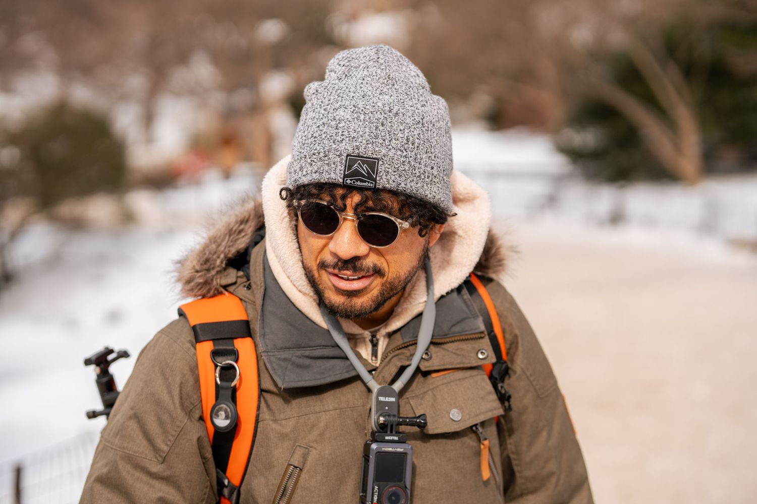 Man wearing grey beanie, sunglasses, and winter jacket with orange backpack outdoors in snow.