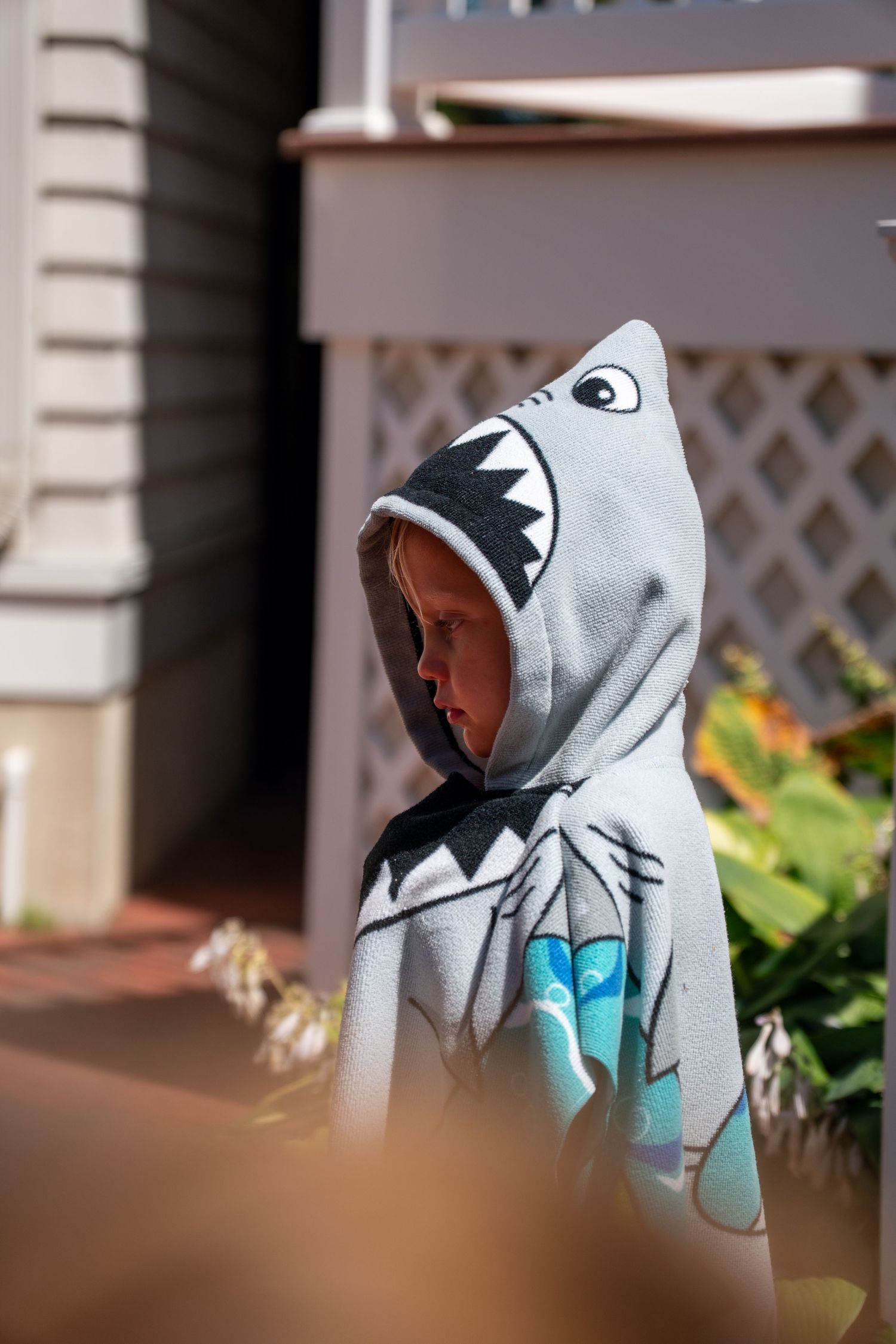 Child in a gray shark hooded towel poncho standing near a house on a sunny day.