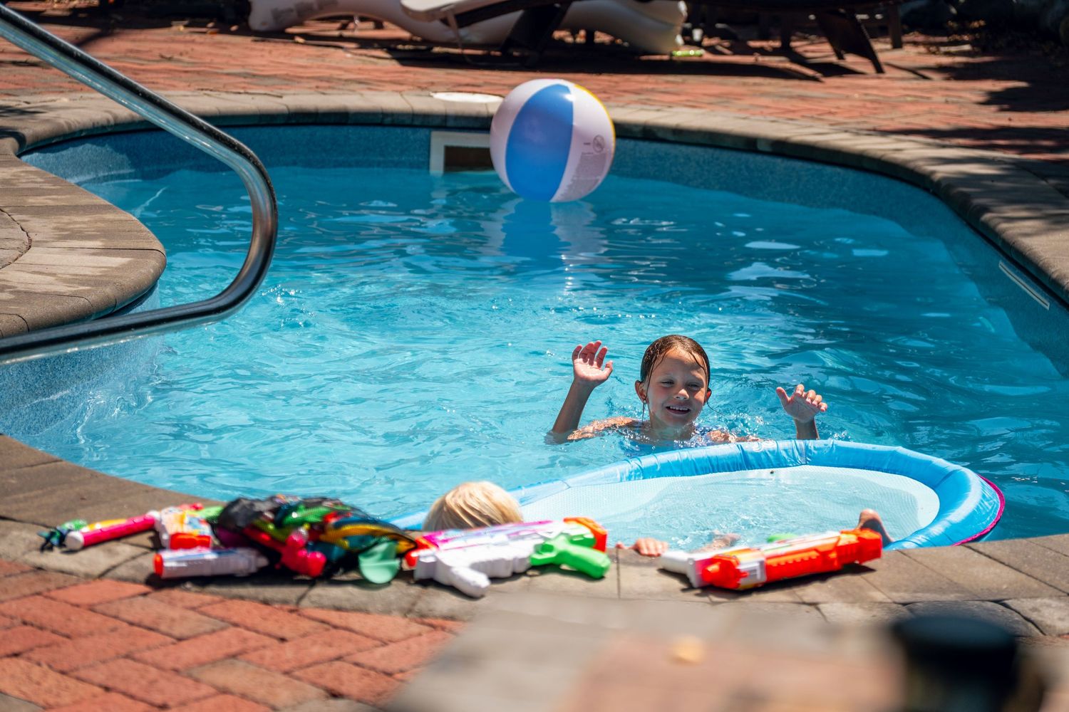 Young child playing in a backyard pool, tossing a beach ball with water toys nearby.