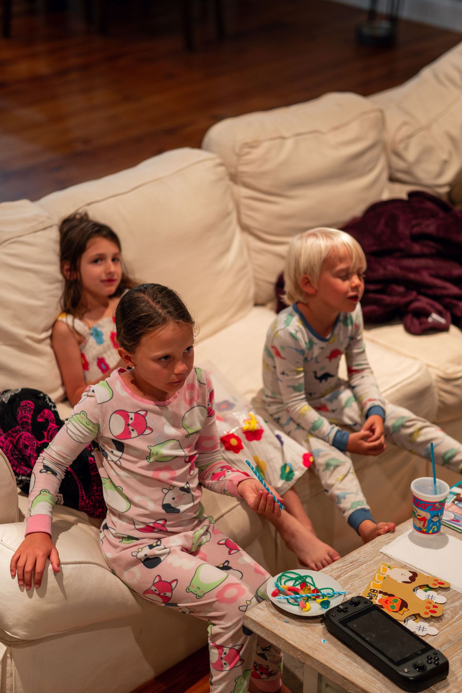 Three kids in pajamas relaxing on a white couch with a Nintendo Switch on the table.