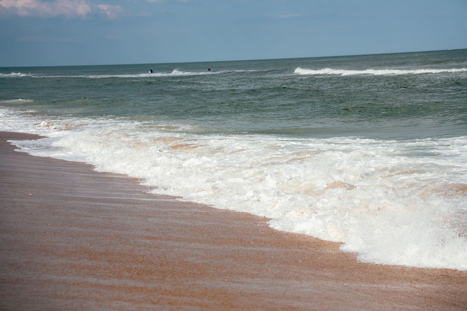 Gentle ocean waves washing onto a sandy beach shore with calm blue-green water and cloudy sky in the background.
