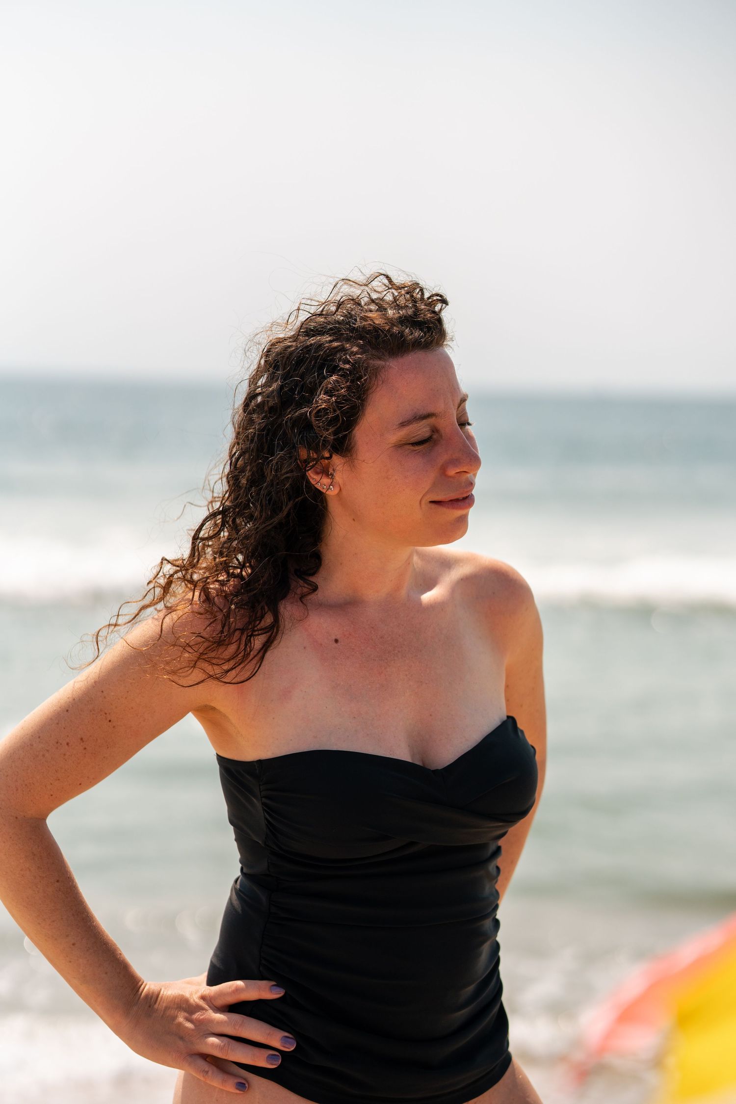 Woman in black strapless swimsuit posing on a sunny beach with ocean waves in the background.