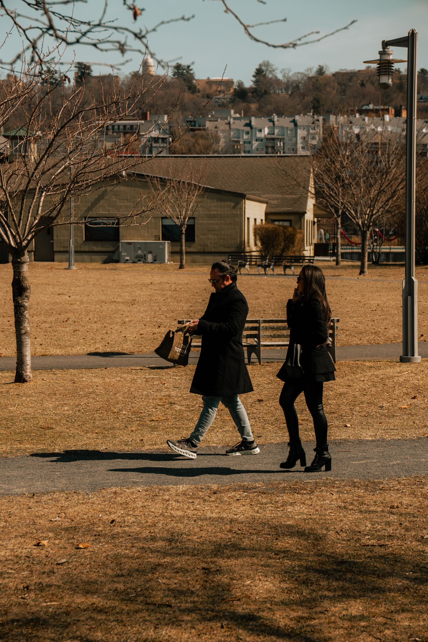 Two people in black coats walking through a bare winter park with trees and buildings in the background.