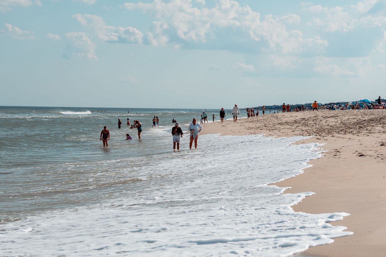 Crowds of beachgoers wading and walking along a wide sandy ocean beach with waves and blue sky.